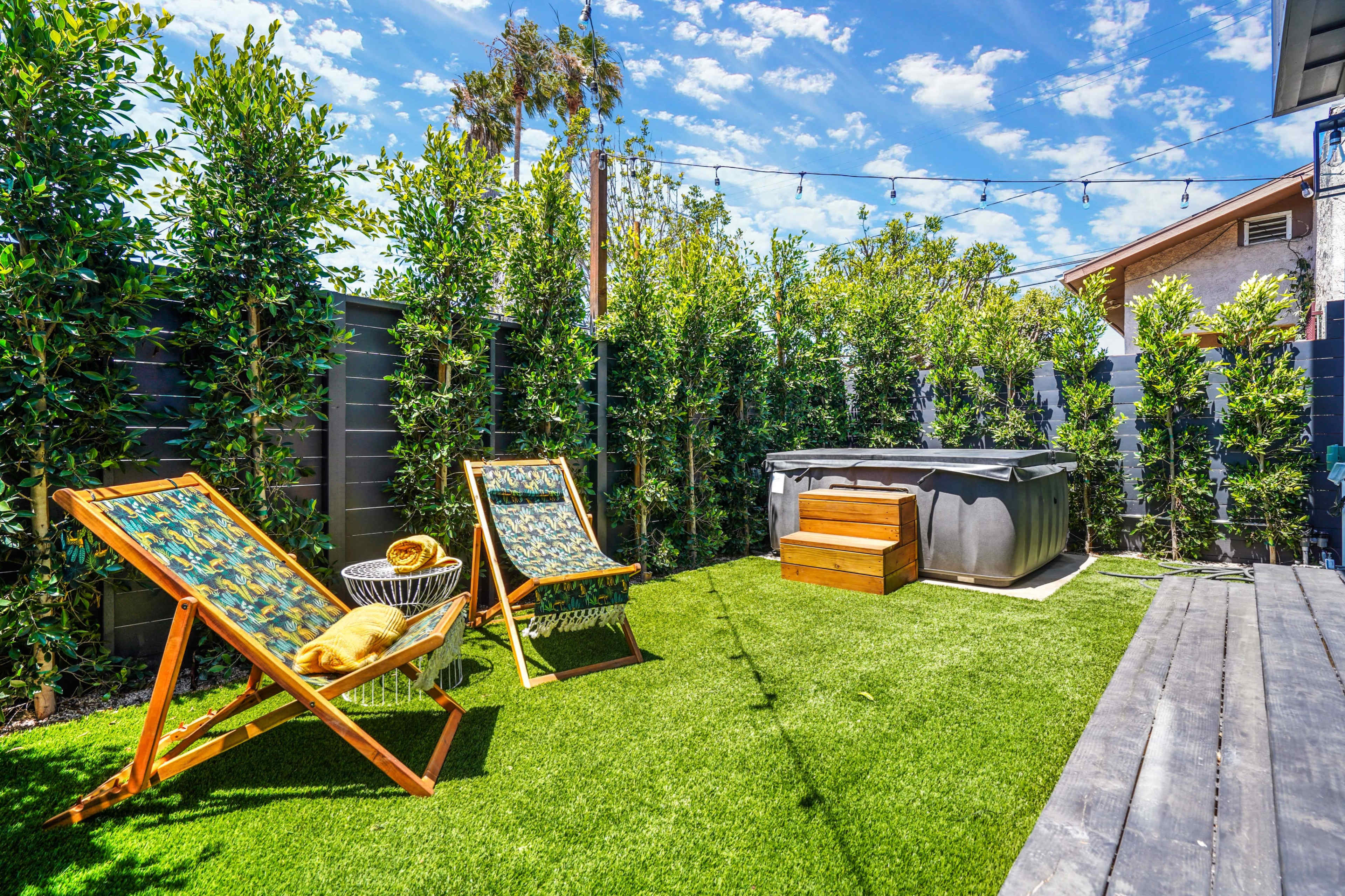 A backyard with two lounge chairs, a hot tub, and lush greenery under a blue sky with scattered clouds.