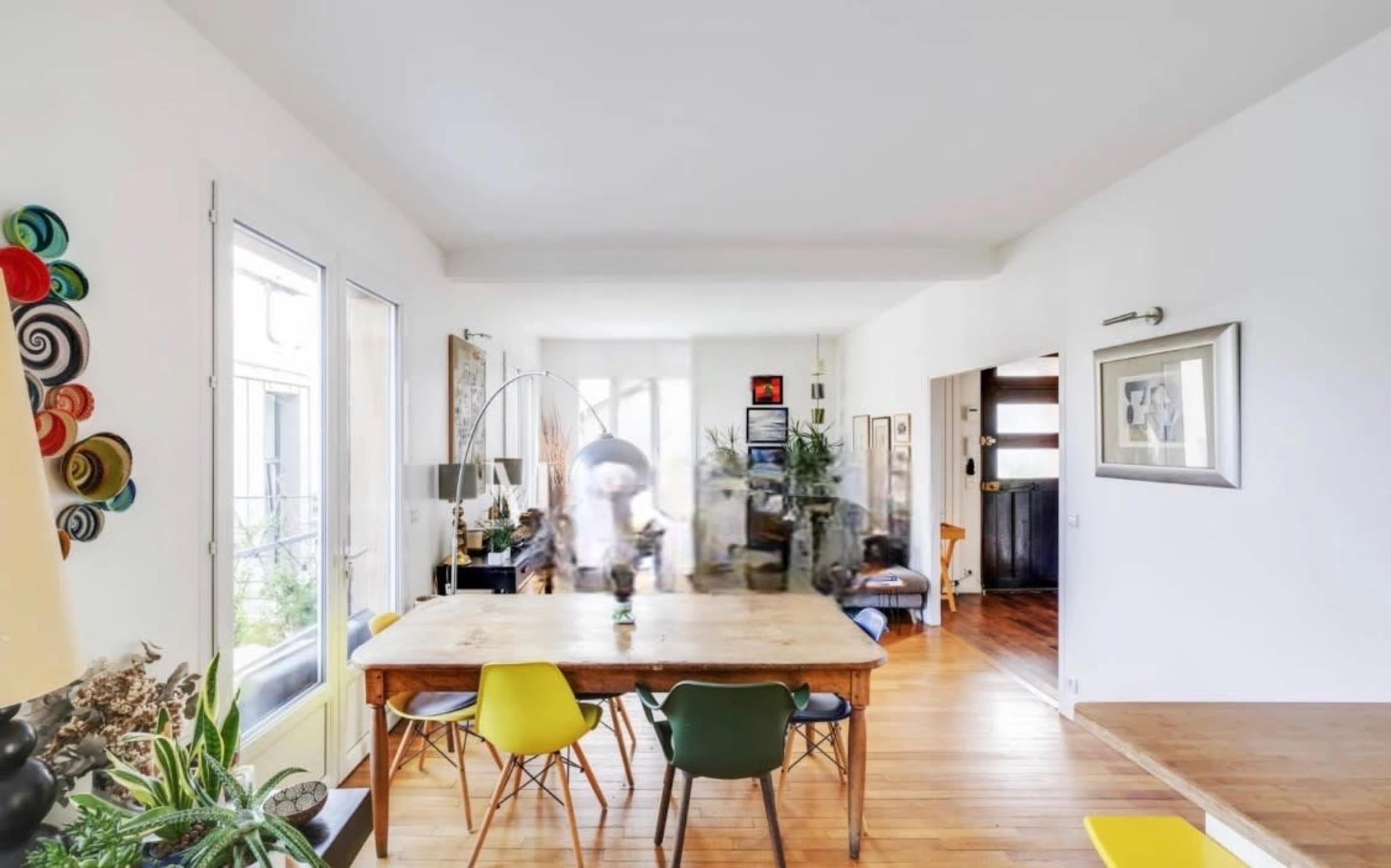 The image shows a bright dining area featuring a large wooden table surrounded by colorful chairs, with large windows allowing natural light to fill the space.