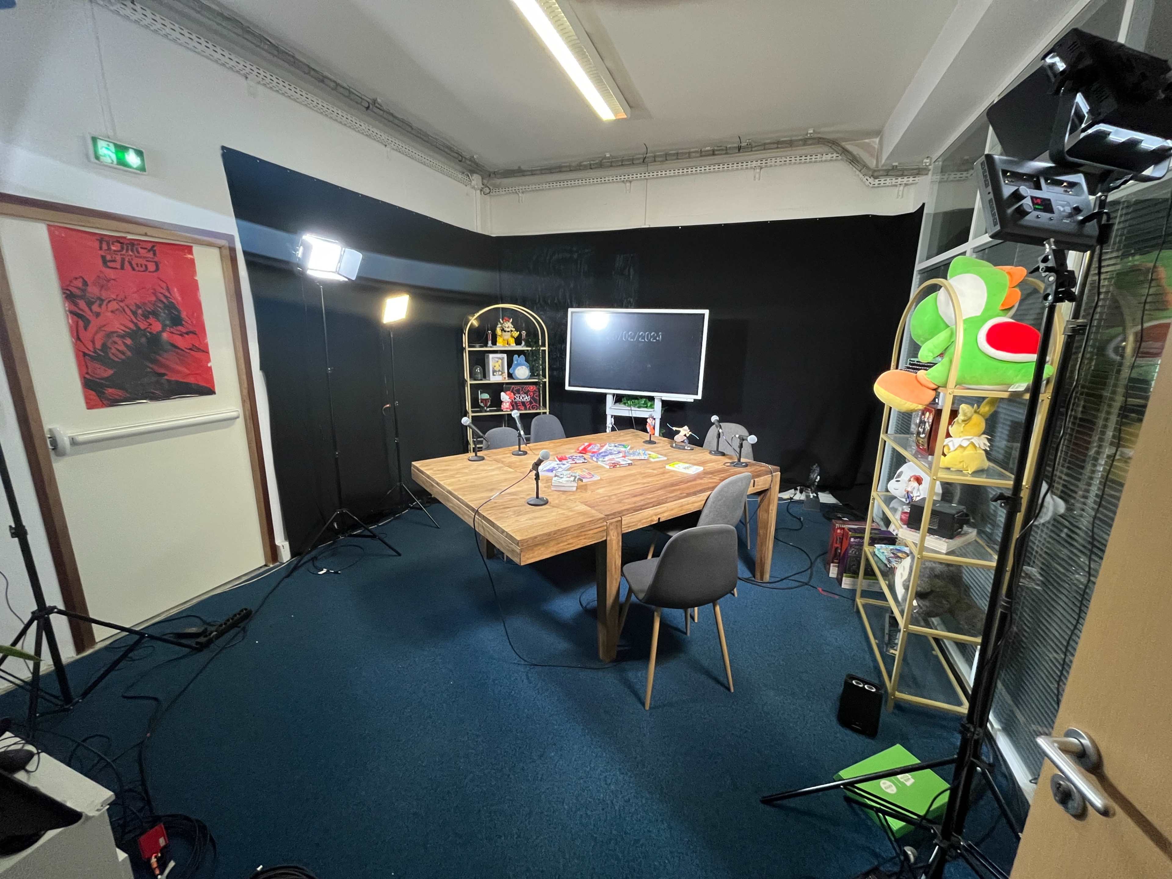 A podcast studio setup with a large wooden table, microphones, a screen, and studio lighting in a room with a black backdrop.