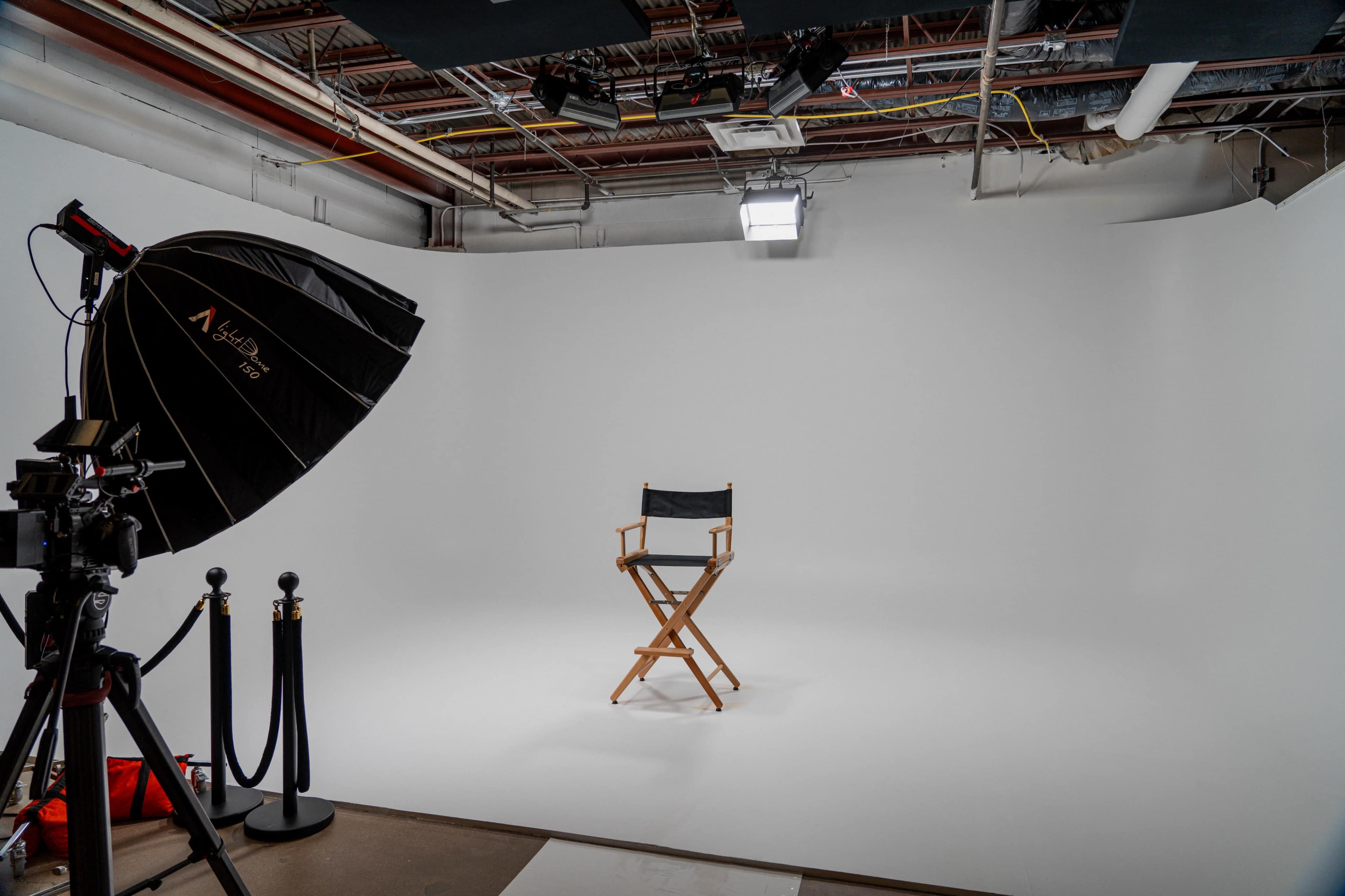 A photography studio features a black director's chair centered in front of a white backdrop, with lighting equipment and a camera positioned nearby.