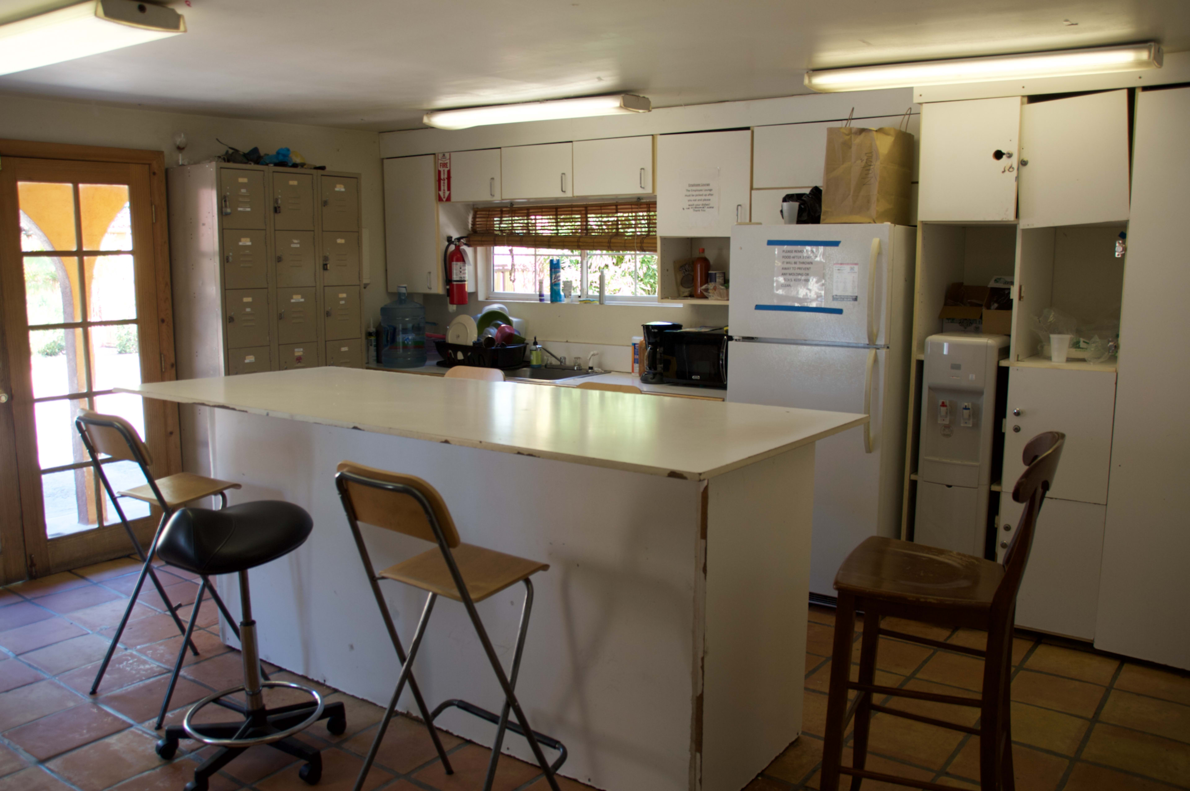 The image shows a simple kitchen with a bar counter, three stools, a refrigerator, and storage cabinets lining the walls.