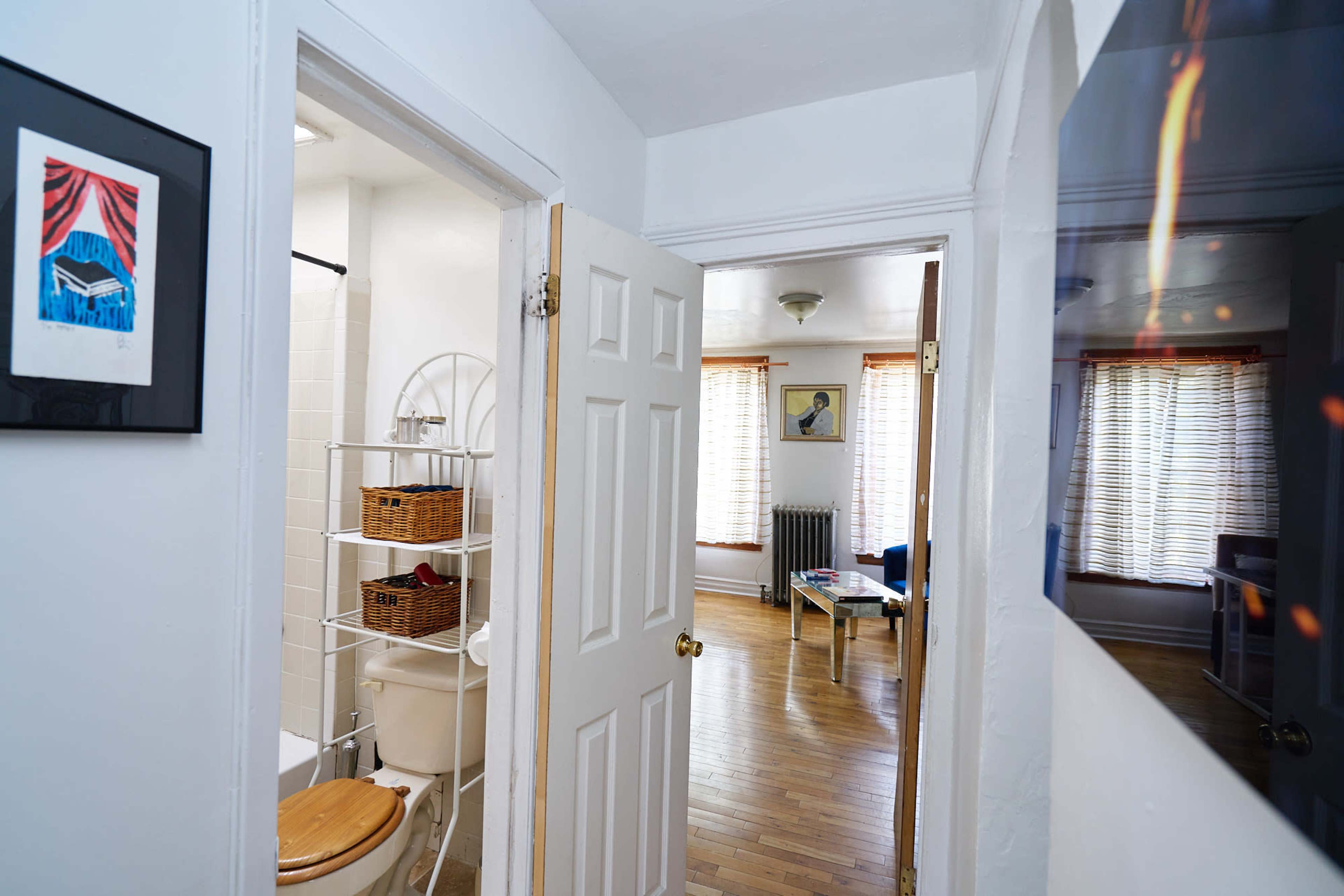 The image shows a hallway leading to a bathroom on the left and a living area on the right, with natural light streaming through window blinds.