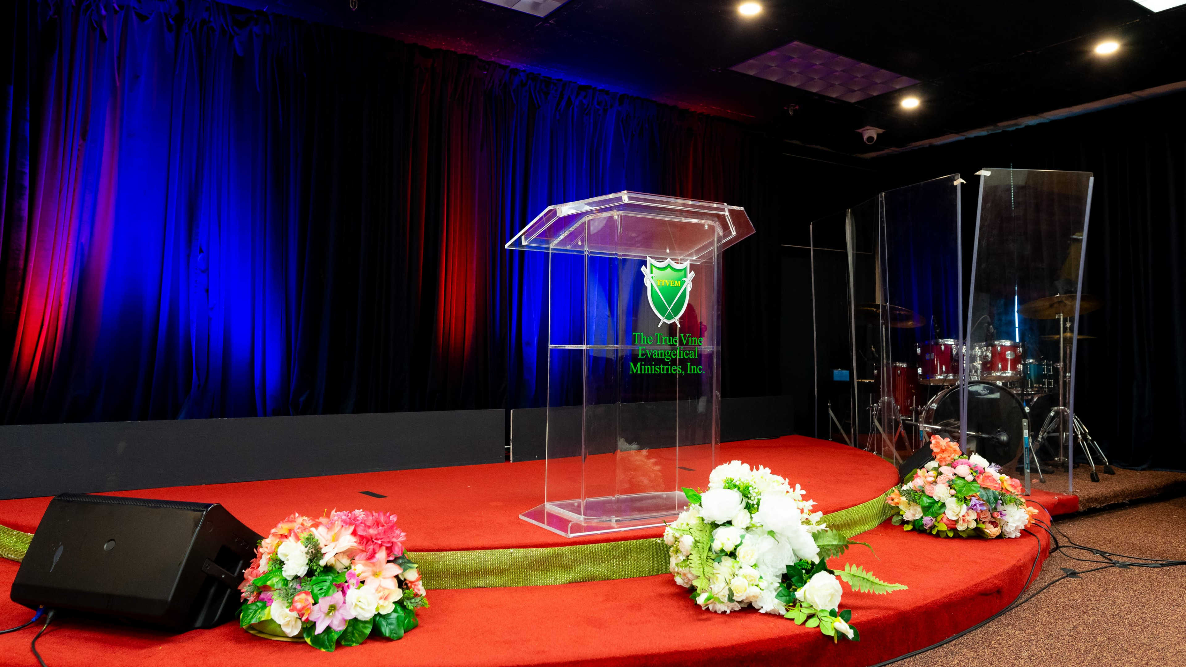 A transparent podium is set on a red carpeted stage in front of a drum set and surrounded by floral arrangements.