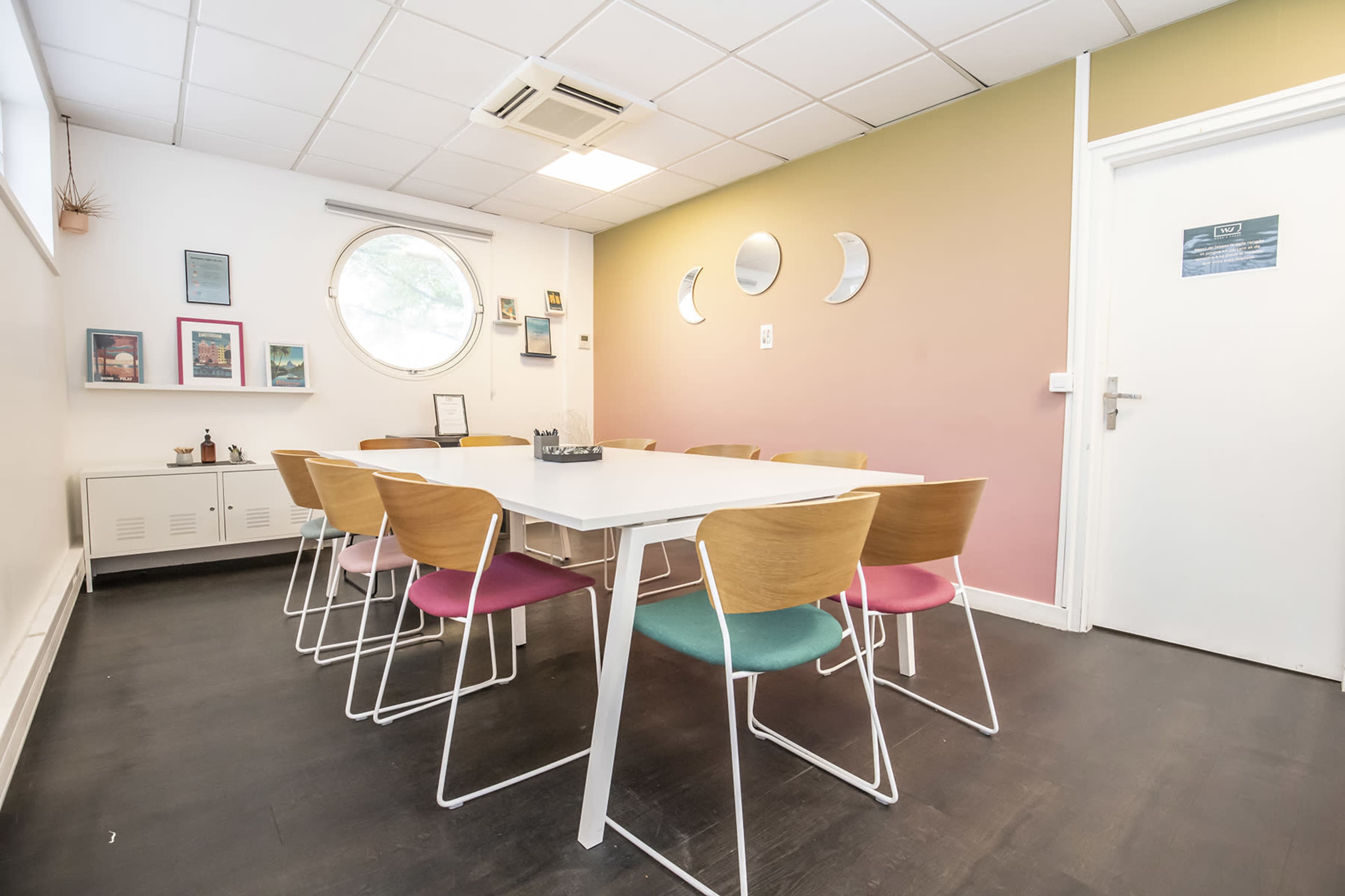 A minimalist meeting room features a white table surrounded by wooden chairs with colored cushions, positioned against a partially painted wall with circular mirrors.