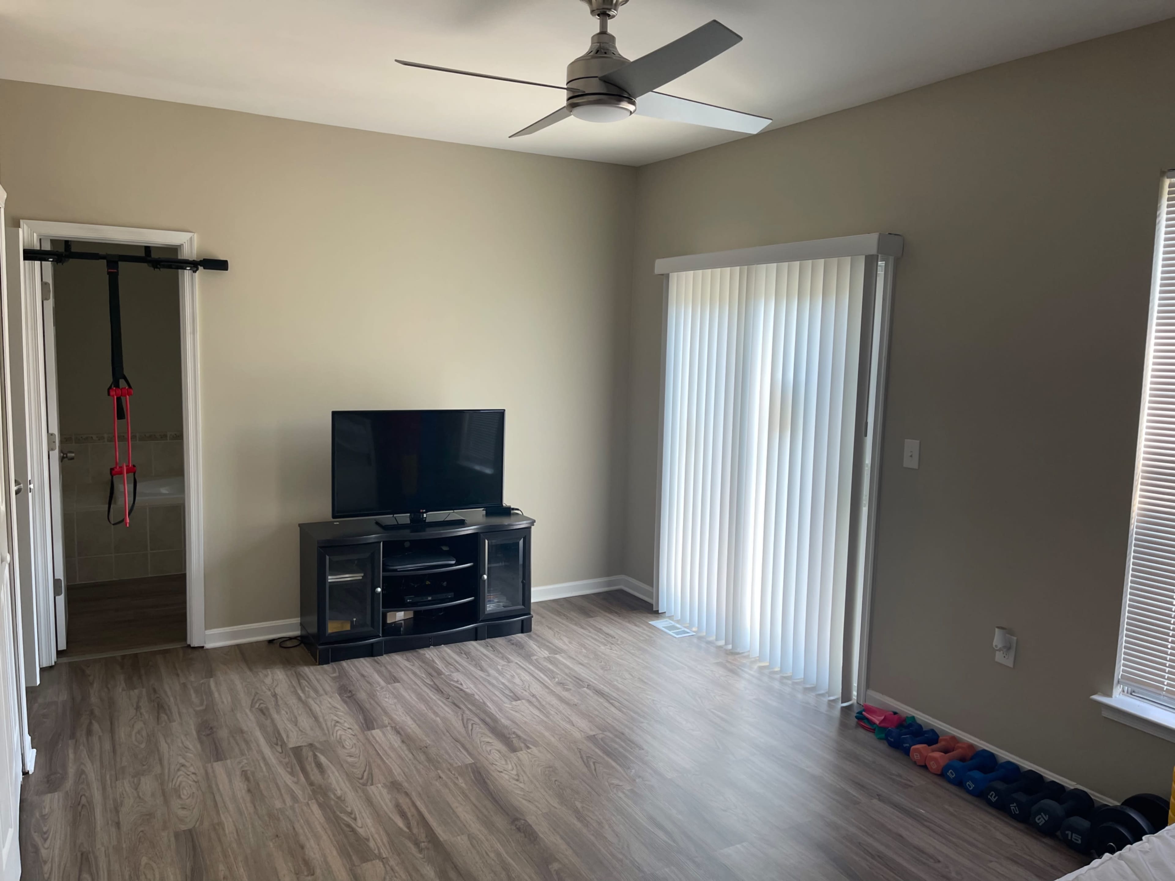 The image shows a spacious, minimally furnished room with a television on a cabinet, a window with vertical blinds, and exercise equipment on the floor.