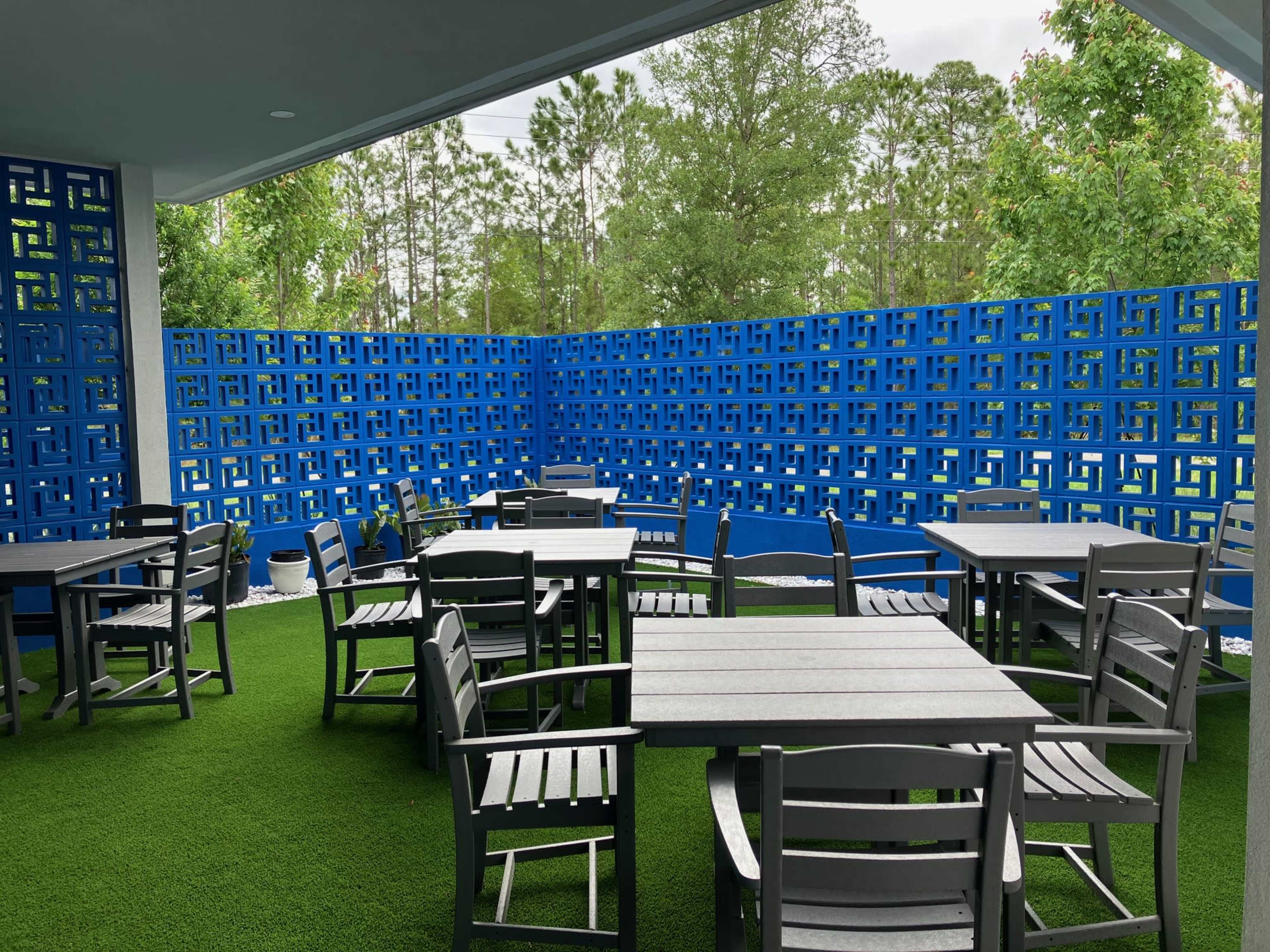 The image shows an outdoor seating area with several gray tables and chairs arranged on artificial green grass, surrounded by a bright blue decorative wall.