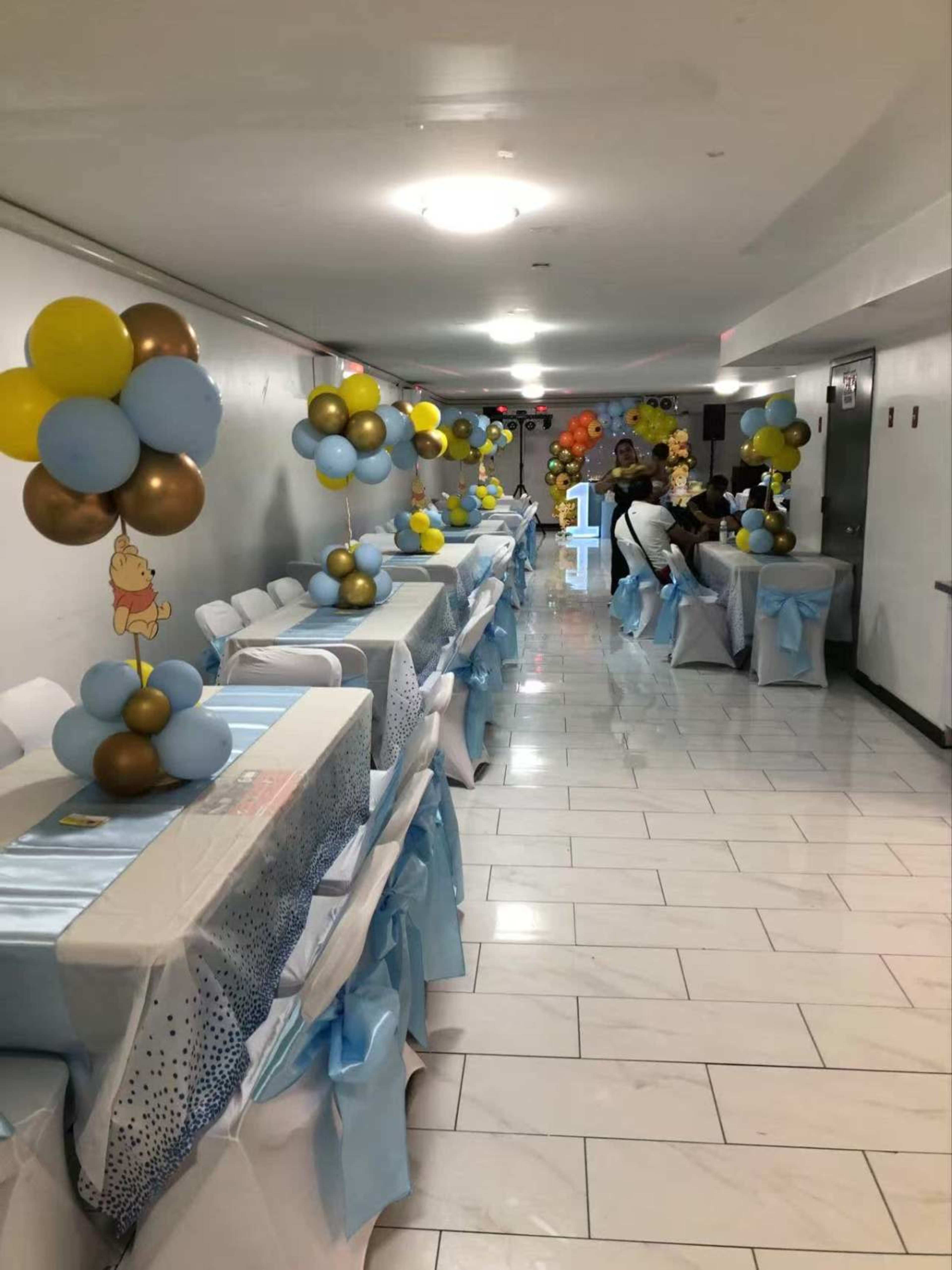 A long hallway is decorated for a party, featuring tables adorned with blue and gold balloons and pastel tablecloths, while guests gather towards the end.