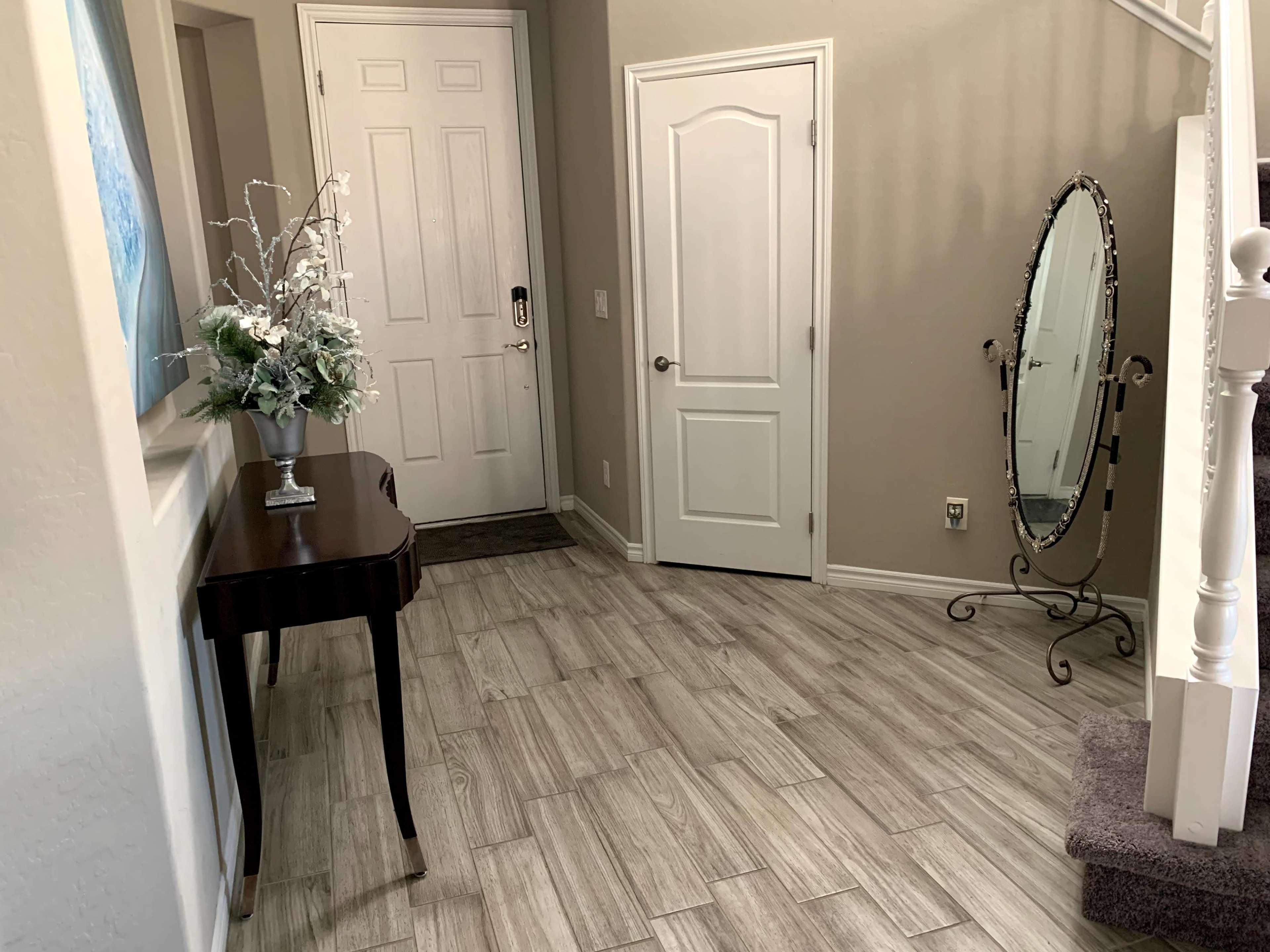 A hallway featuring a small table with a vase, a door leading outside, a closed interior door, a staircase, and a decorative mirror mounted on a stand.