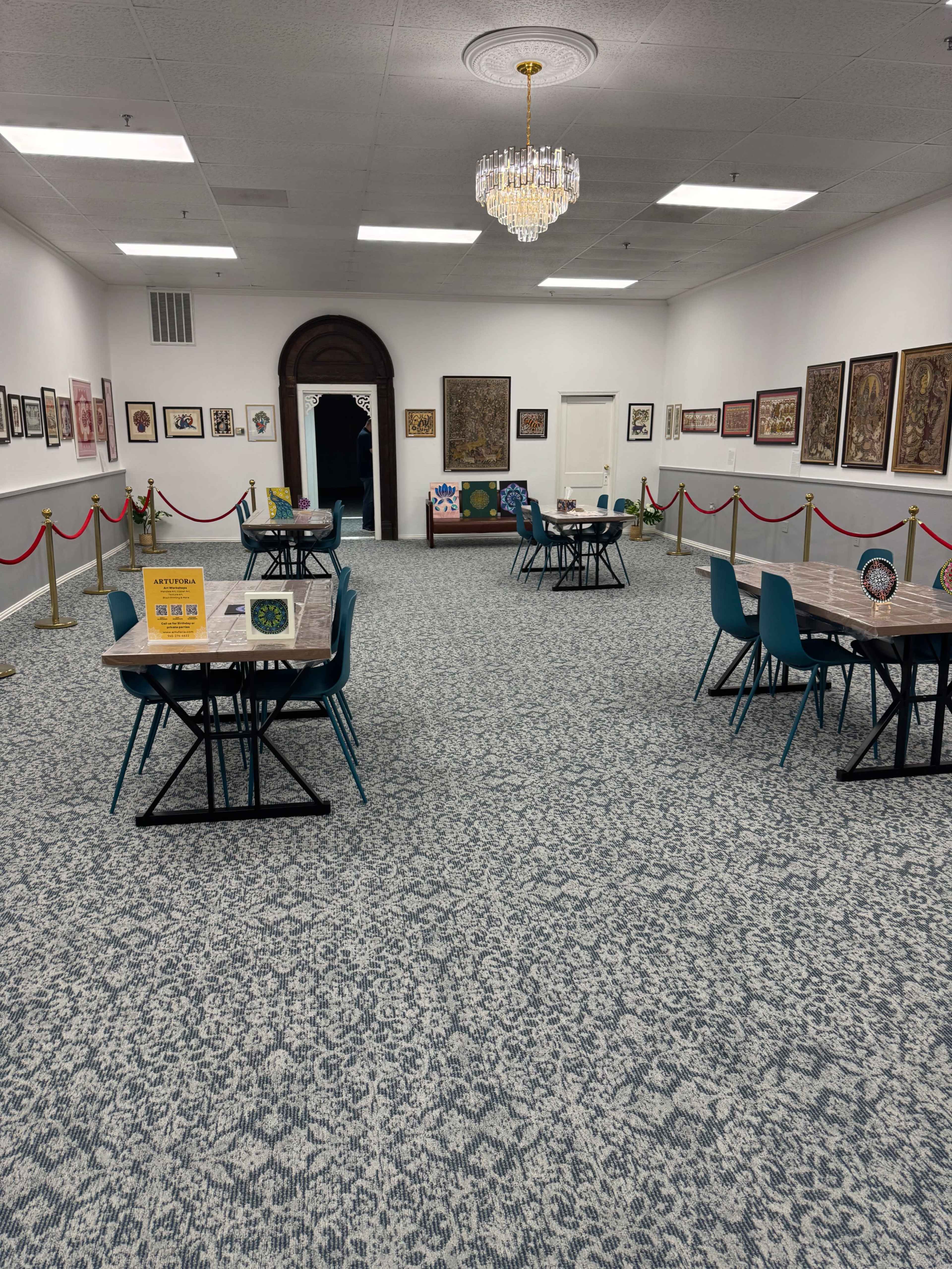 The image shows a spacious gallery or room featuring multiple tables with chairs, artwork displayed on the walls, and a chandelier above.