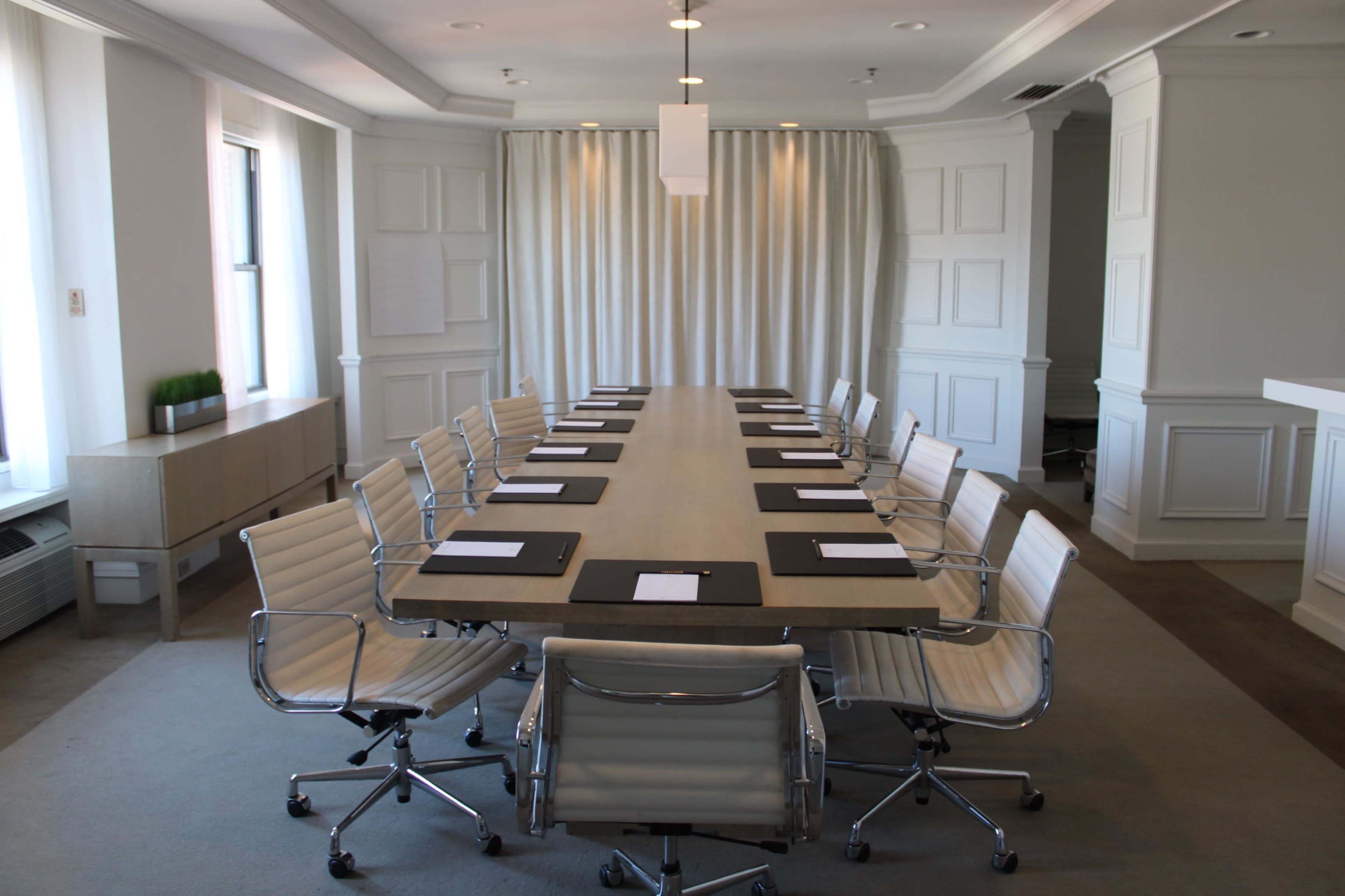 A long conference table with chairs is arranged in a brightly lit meeting room, featuring white walls and a curtain backdrop.