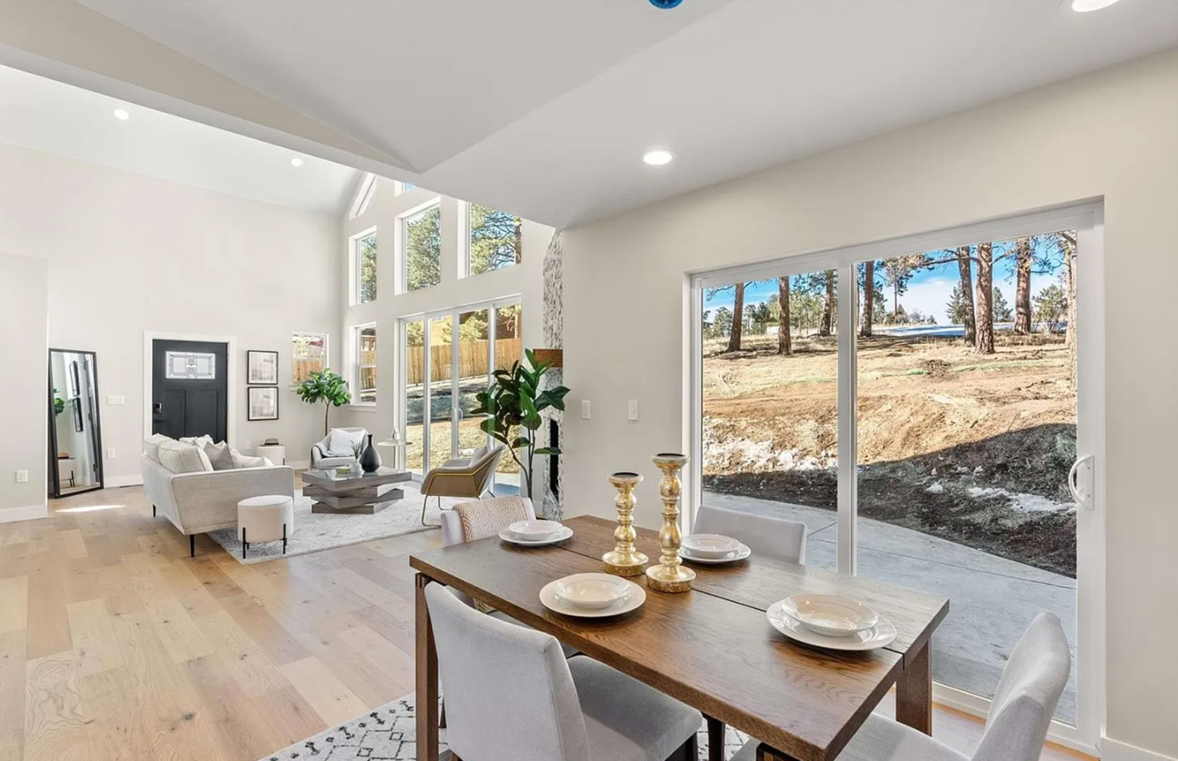 A modern dining area features a wooden table set for four, with a spacious living room visible in the background and large windows offering views of a tree-lined outdoor space.