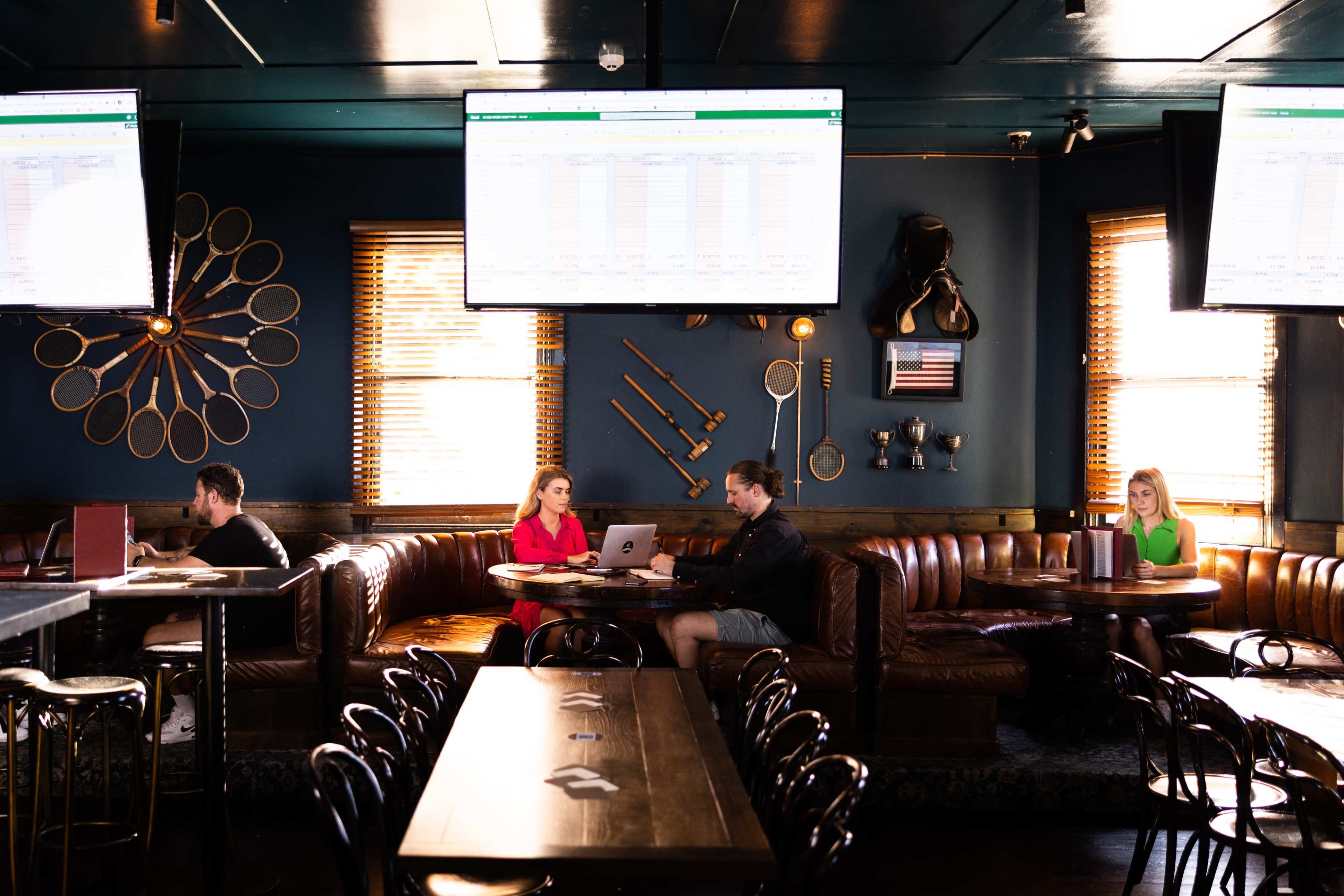 A restaurant interior features dark walls, leather seating, and large screens displaying data, with three patrons engaged in work and conversation.