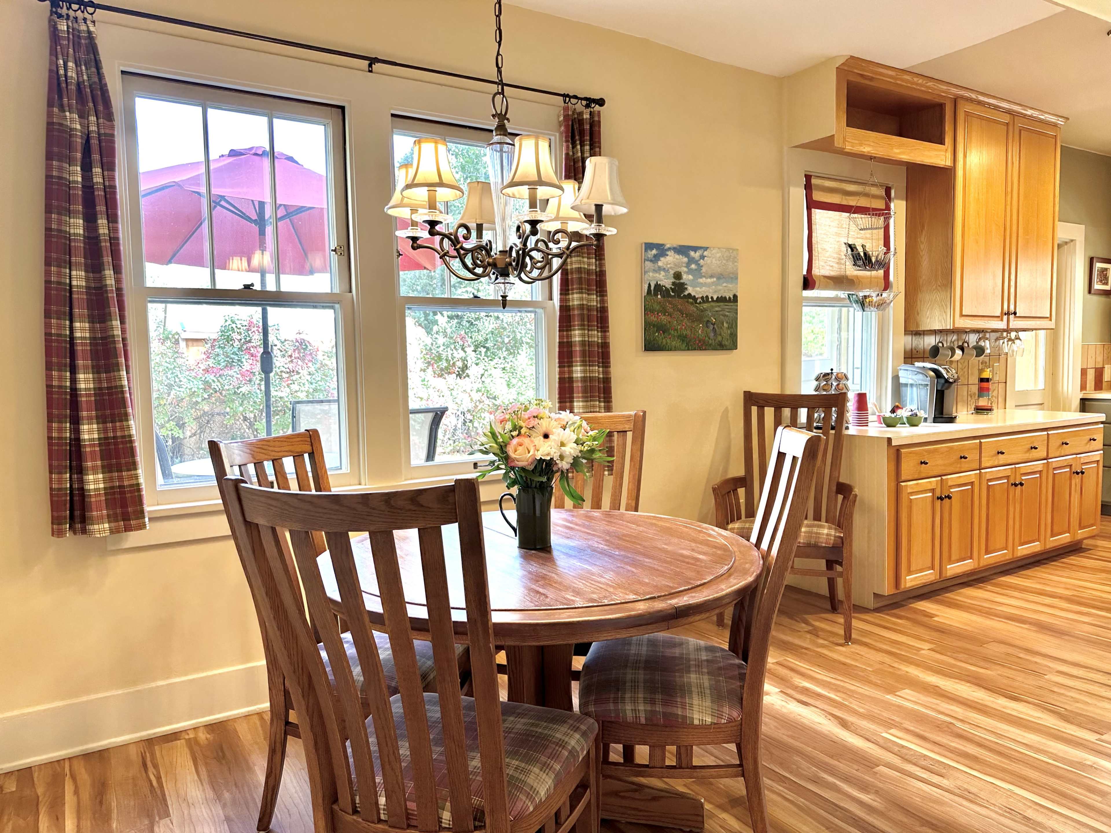 A cozy dining area features a round wooden table surrounded by four chairs, a floral centerpiece, and a window with a view of an outdoor patio umbrella.