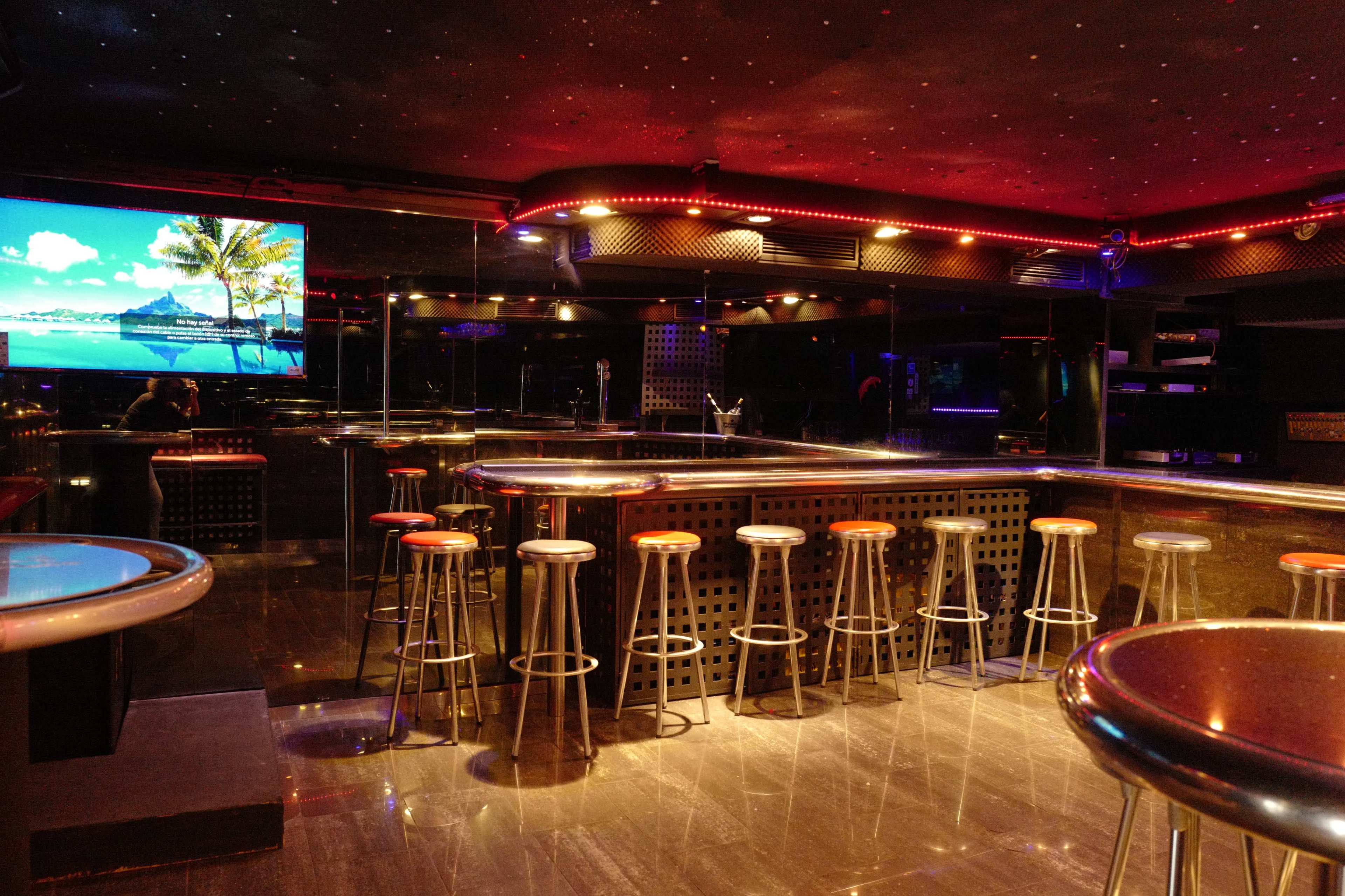 An empty bar area with metal stools and a large screen displaying a scenic view, illuminated by colorful lighting.