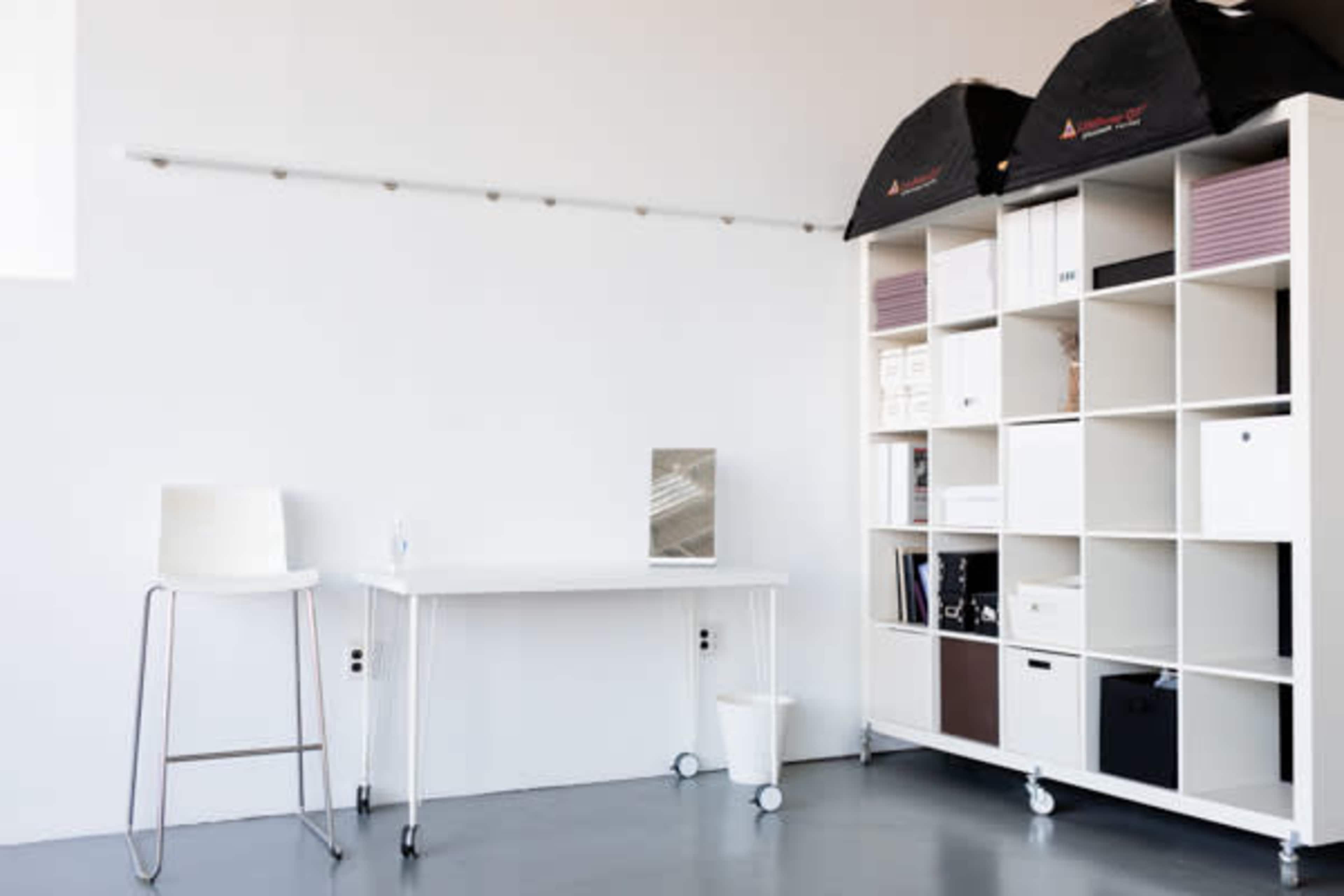 A minimalist office space with a white table on wheels, a stool, and a shelving unit filled with boxes and materials, against a plain wall.