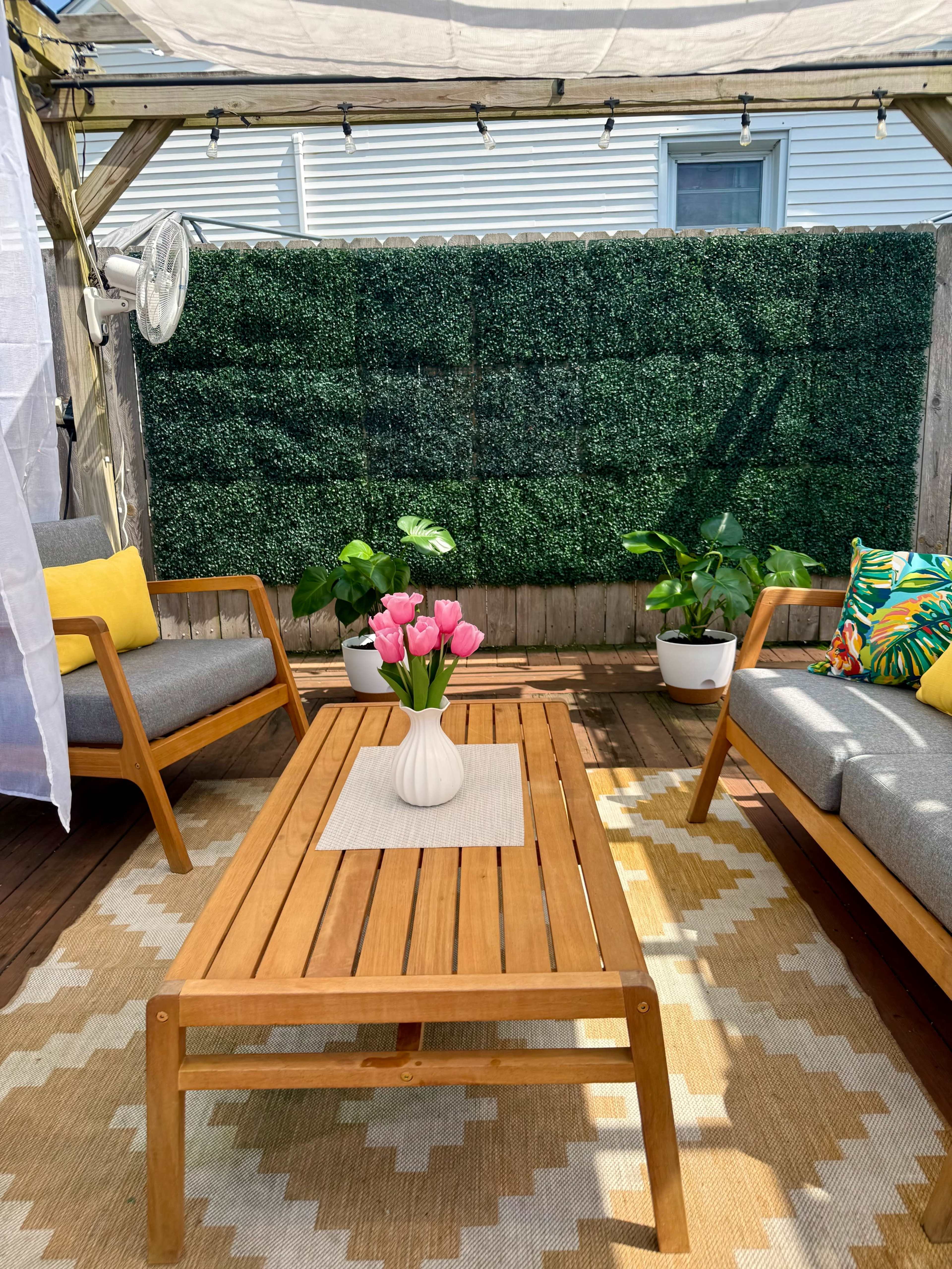 The image shows a cozy outdoor seating area with wooden furniture, a coffee table adorned with a vase of pink flowers, and greenery in the background.