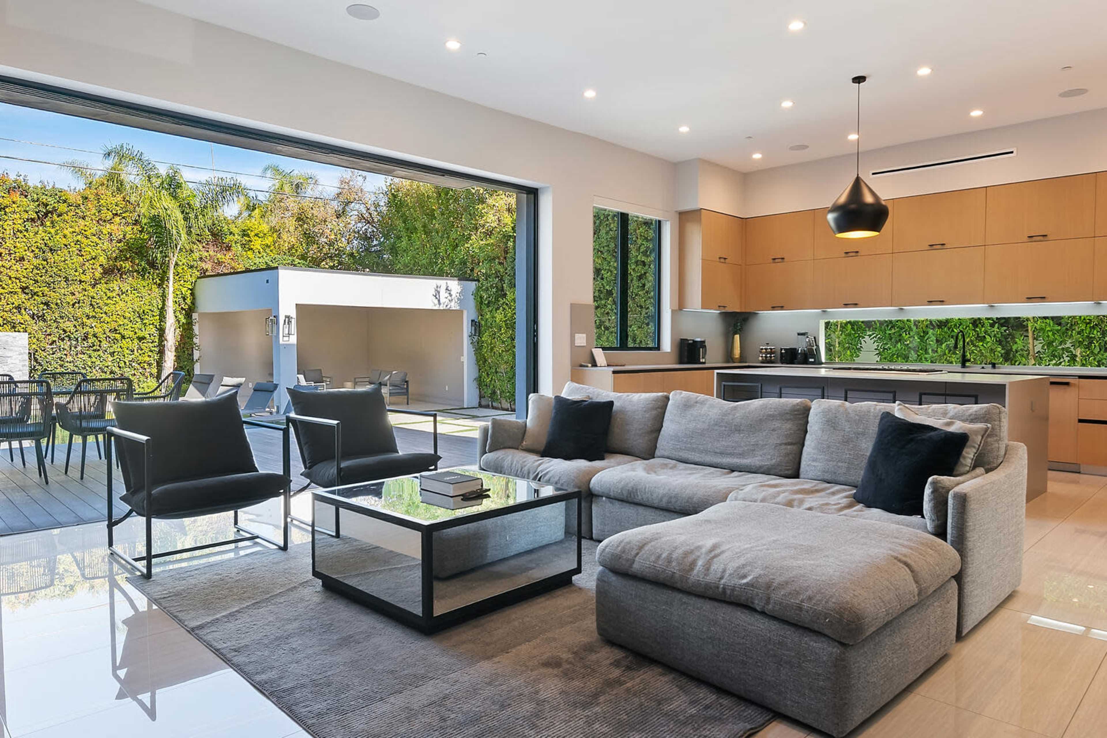 A modern living room features a large sectional sofa, a coffee table, and sliding glass doors that open to a patio with outdoor seating.