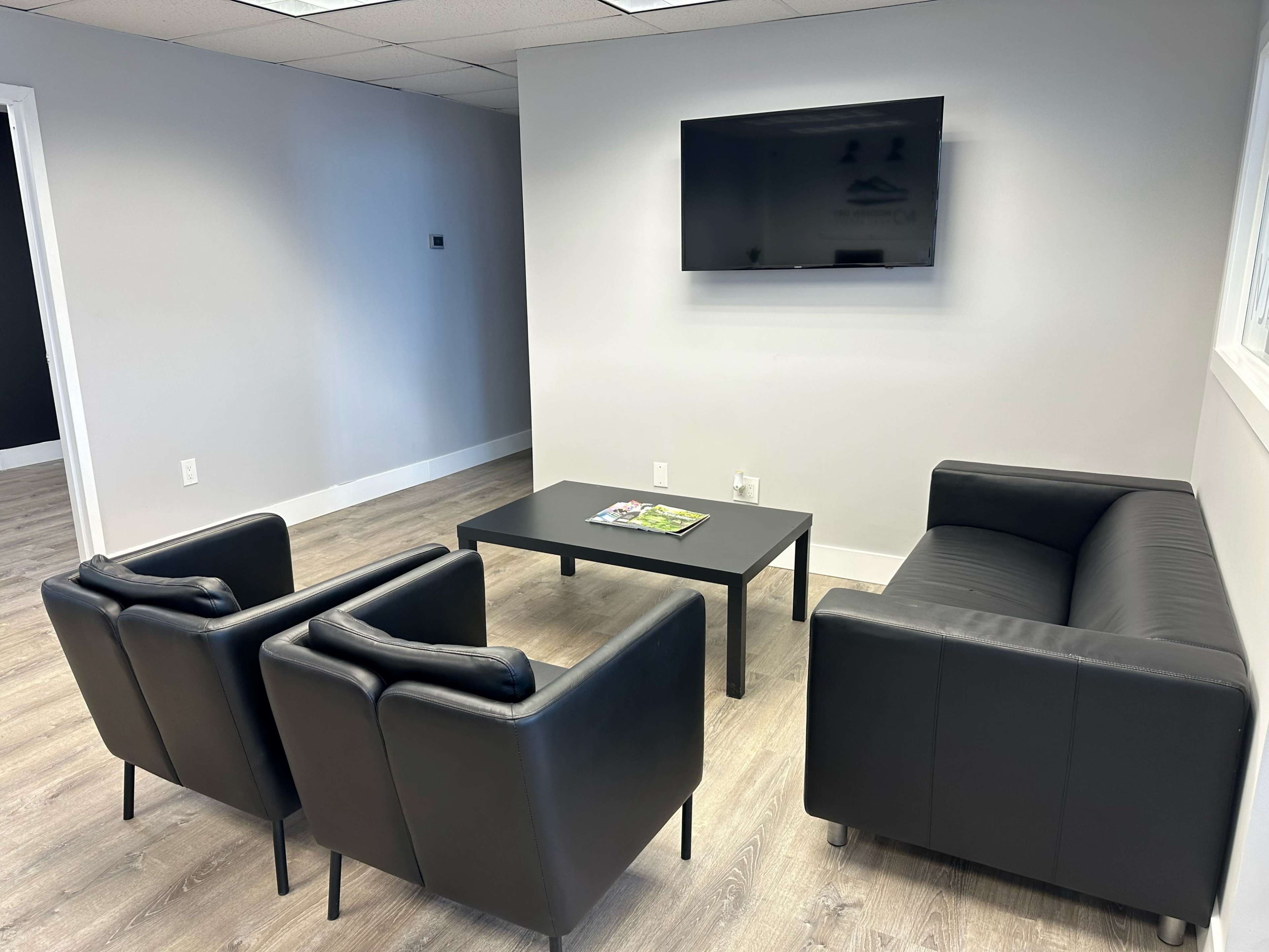 A modern waiting area featuring two black couches, three chairs, a coffee table, and a wall-mounted television.