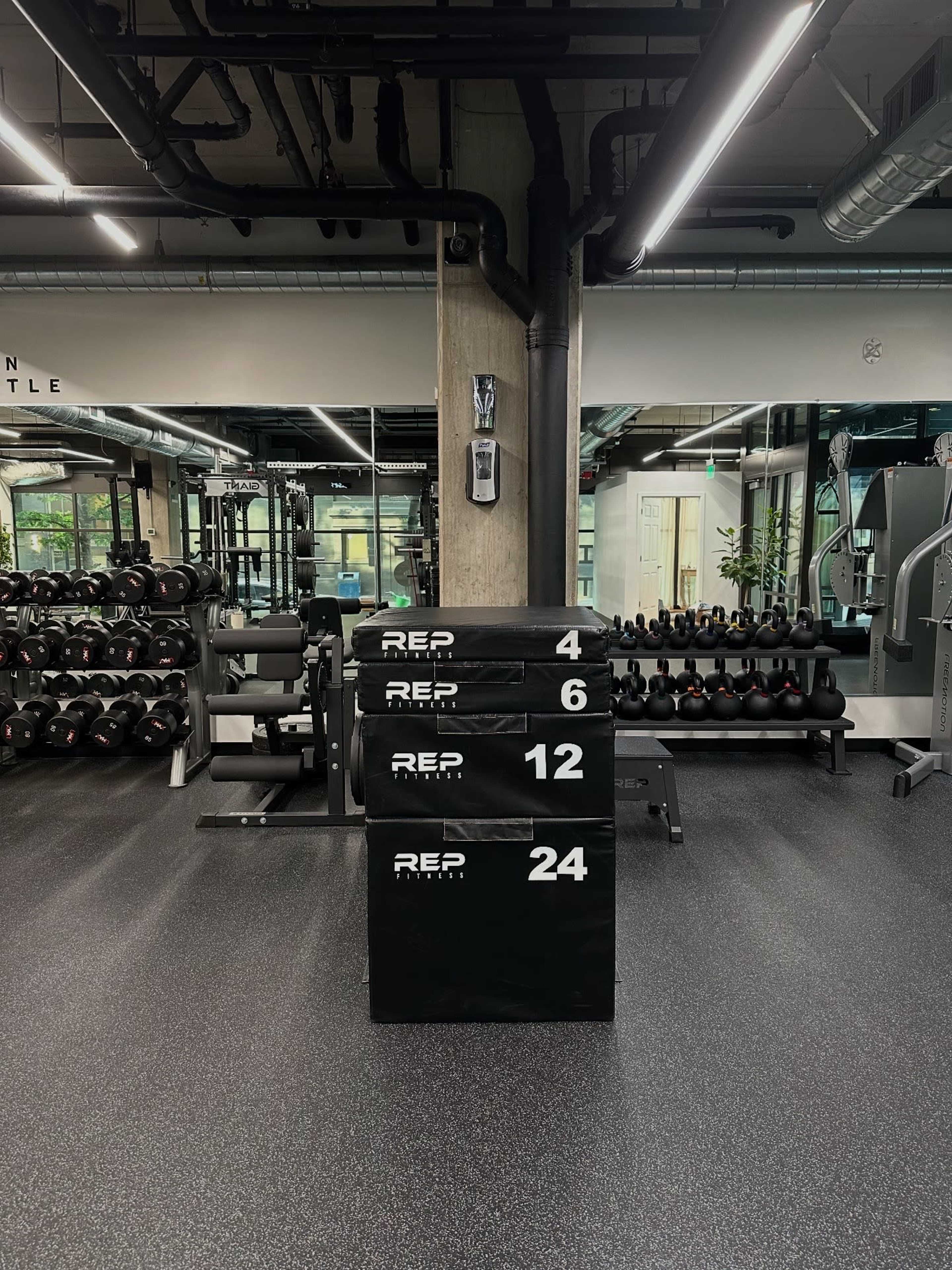 The image shows a gym interior featuring a black stack of foam boxes labeled with numbers, surrounded by various gym equipment and dumbbells.