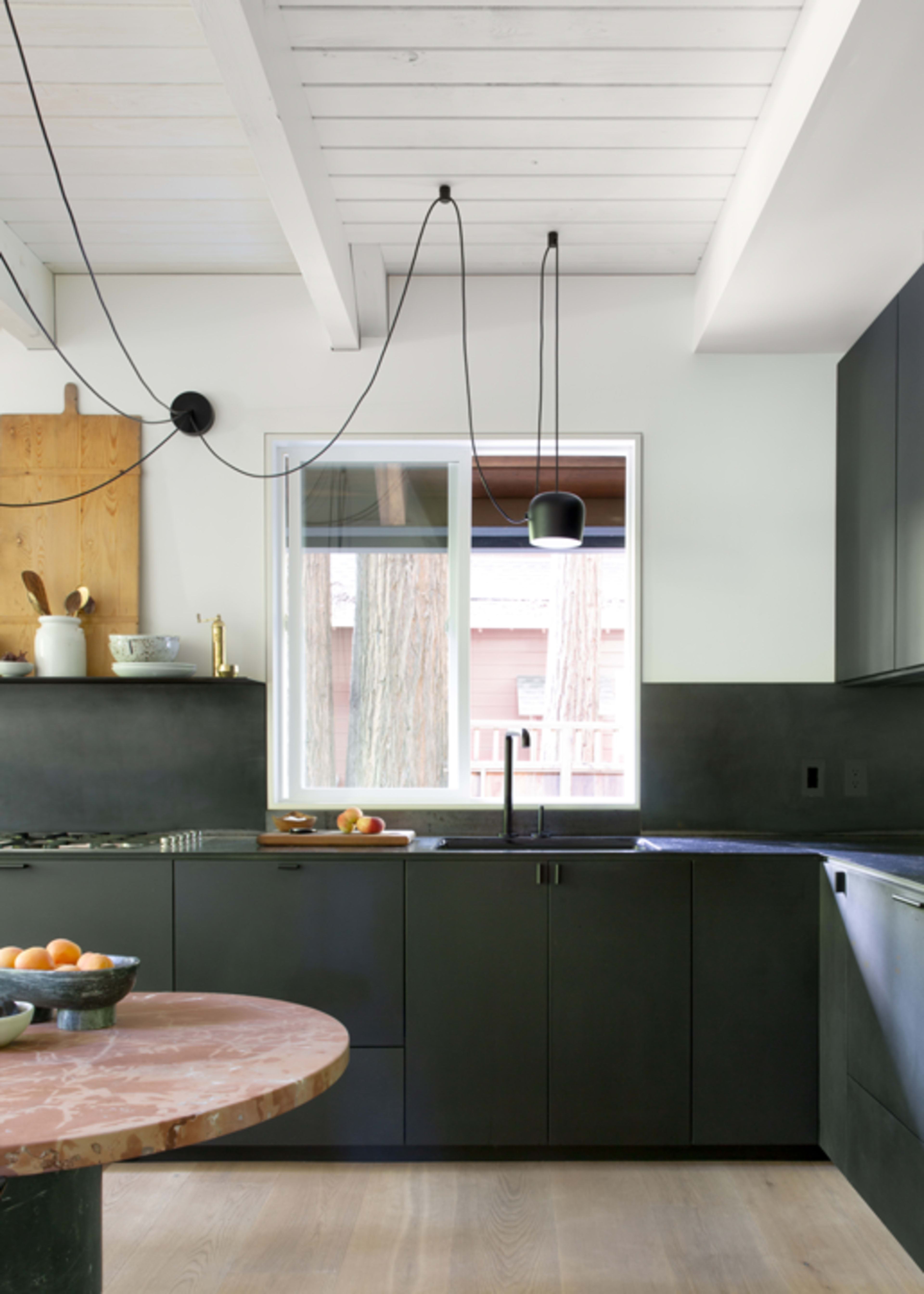 The image shows a modern kitchen featuring black cabinetry, a round marble table, and a large window with a view of trees outside.