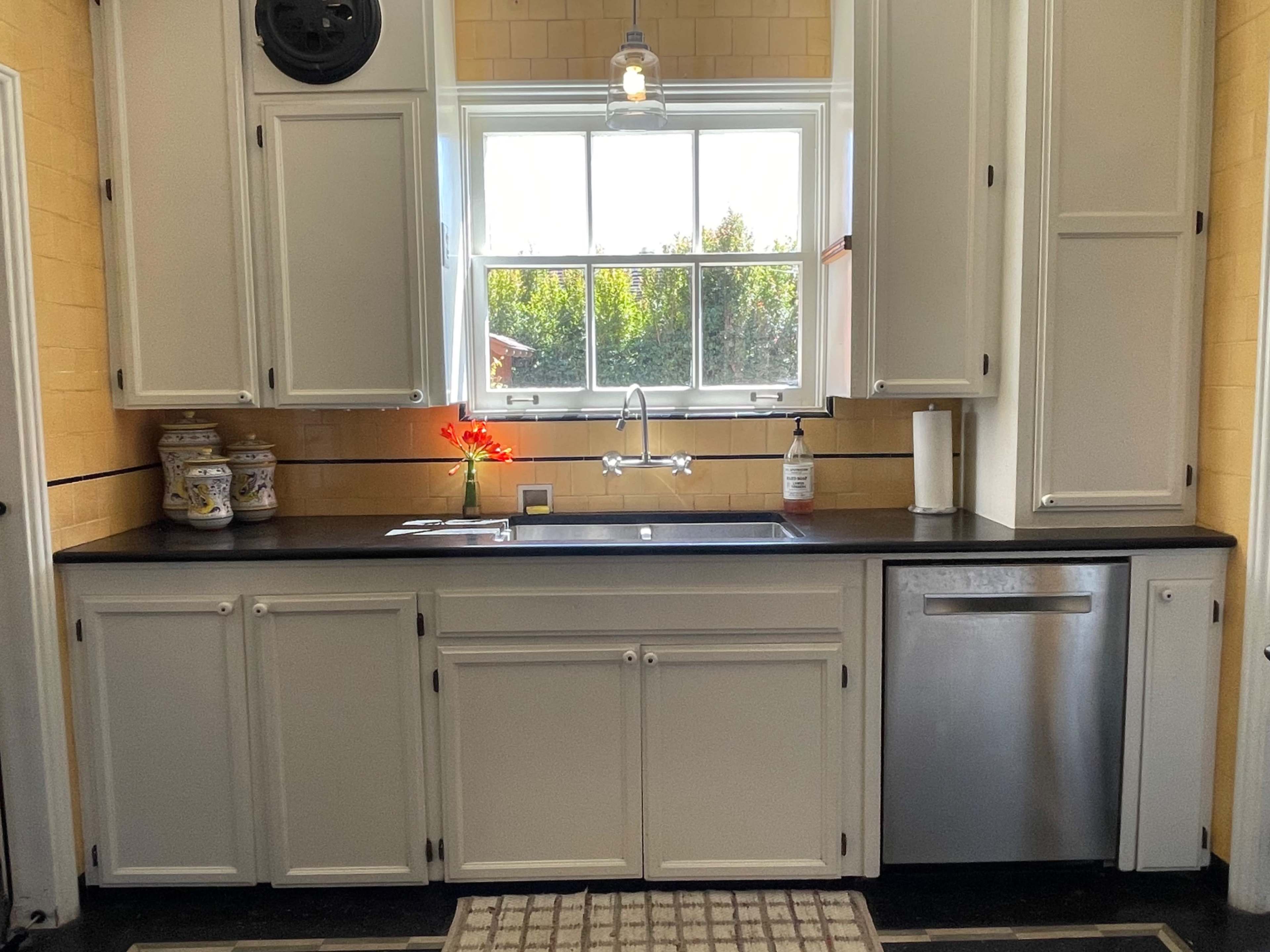 A kitchen with white cabinets, a black countertop, a sink beneath a large window, and a stainless steel dishwasher.