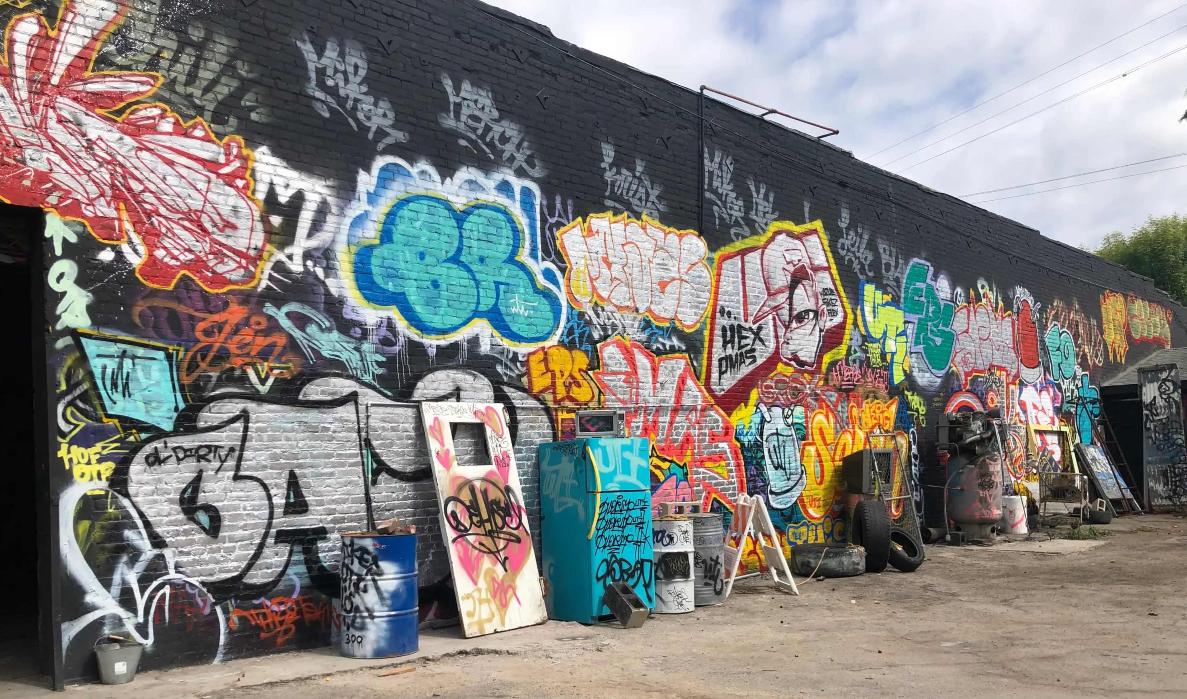 The image shows a colorful wall covered in graffiti art, with various designs and text, alongside a cluttered alley featuring barrels and other debris.