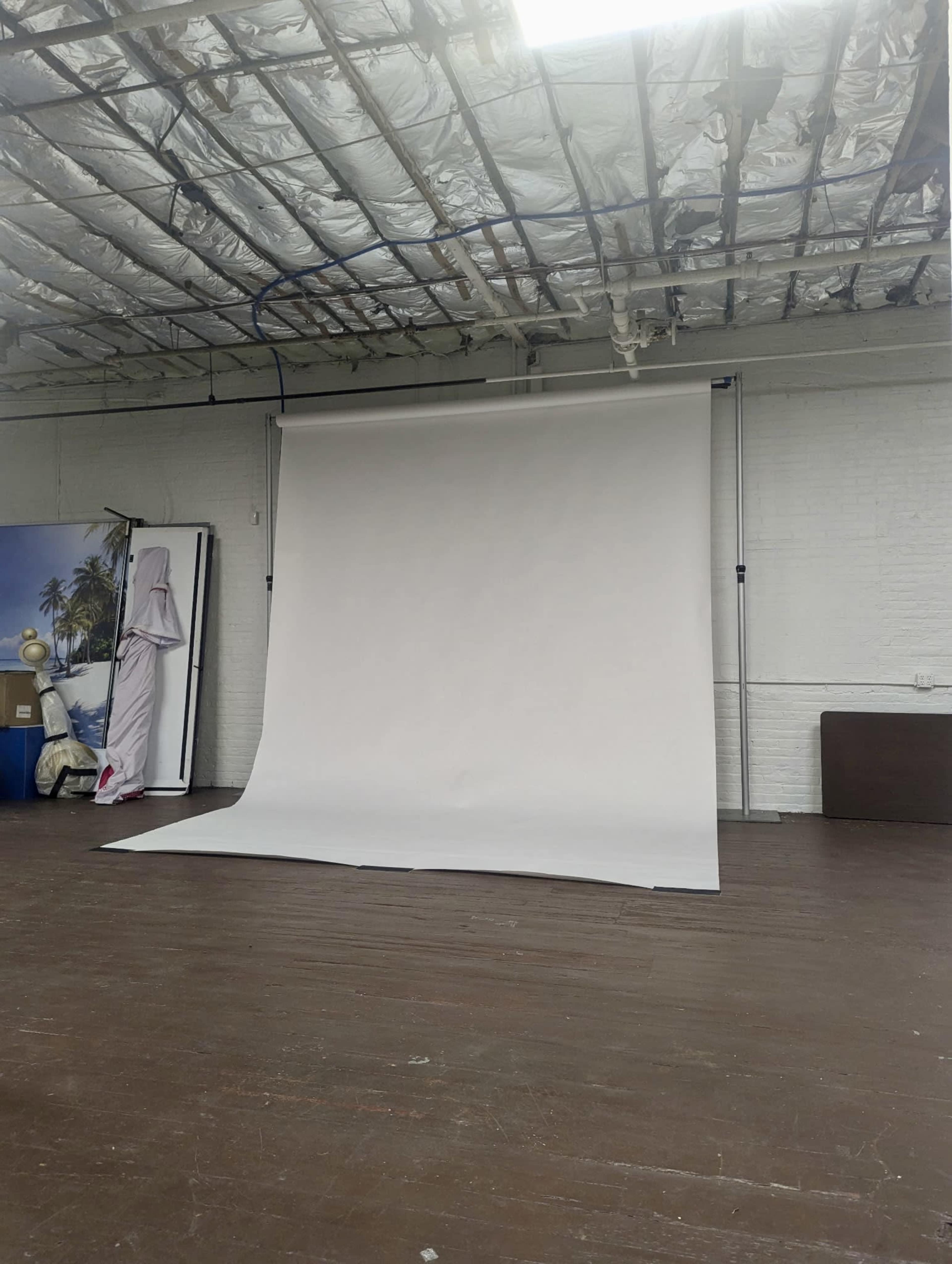 The image shows an empty photography studio with a large white backdrop, wooden flooring, and exposed ceiling beams.