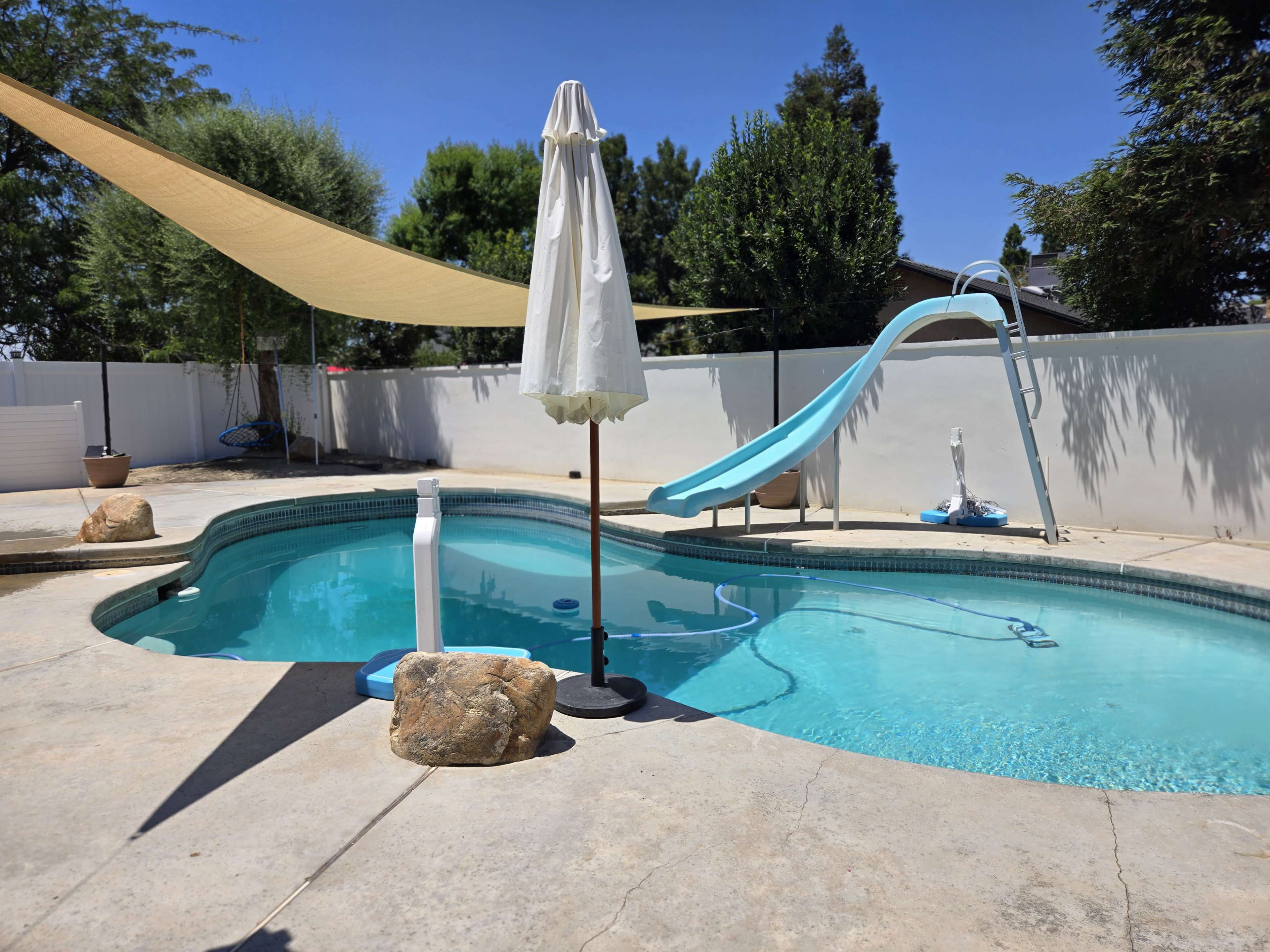 A swimming pool with a slide, an umbrella, and shaded areas created by fabric canopies.