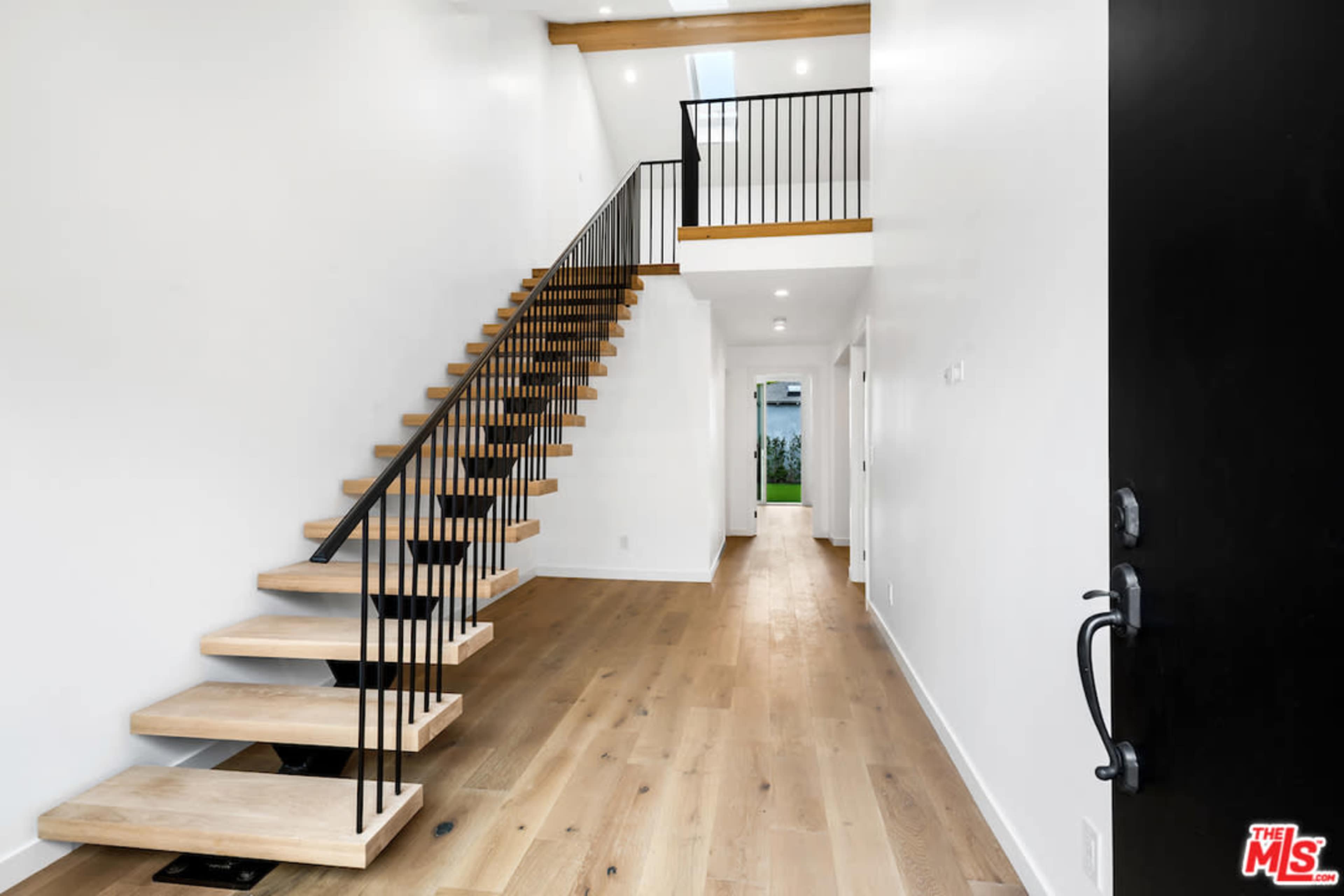 A modern entryway featuring a staircase with wooden steps and black metal railings, leading to a second level, with white walls and wooden flooring.
