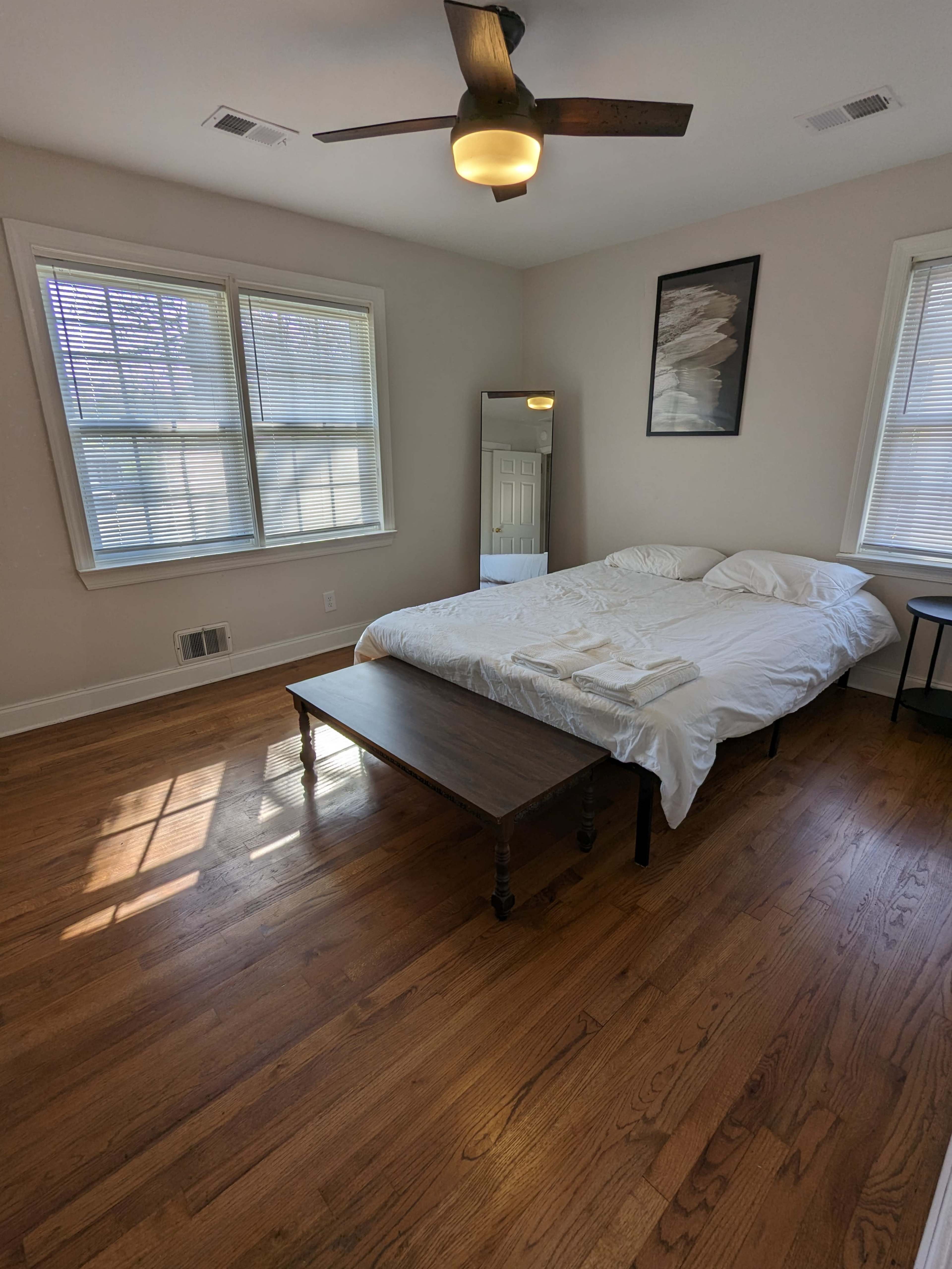 The image shows a well-lit bedroom with a bed, a wooden table, a large mirror, and two windows.