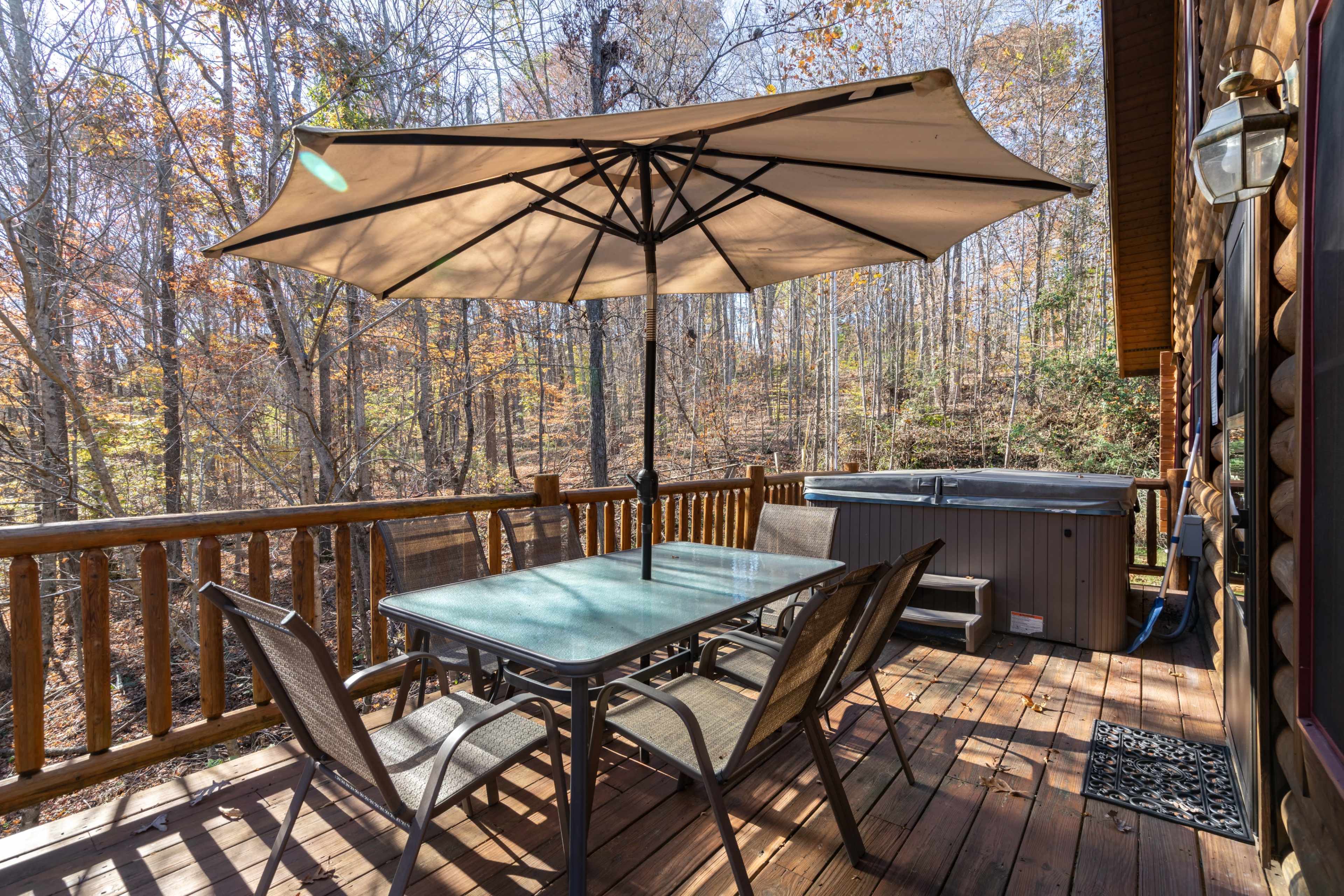 A deck features a large umbrella over a glass table surrounded by chairs, with a hot tub and wooded area in the background.