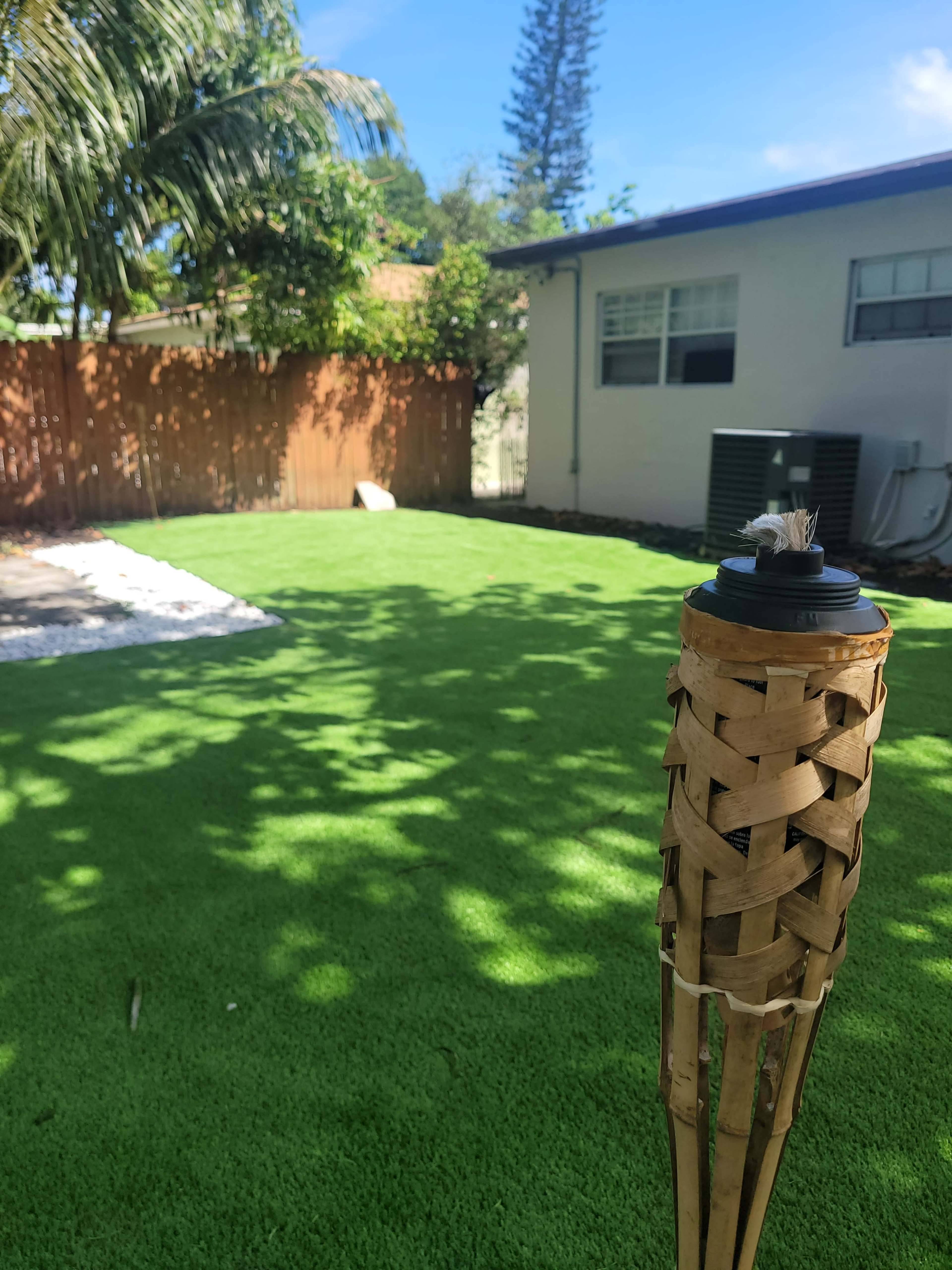 A well-maintained yard featuring artificial green grass, a bamboo tiki torch in the foreground, and a house with a light-colored exterior in the background.