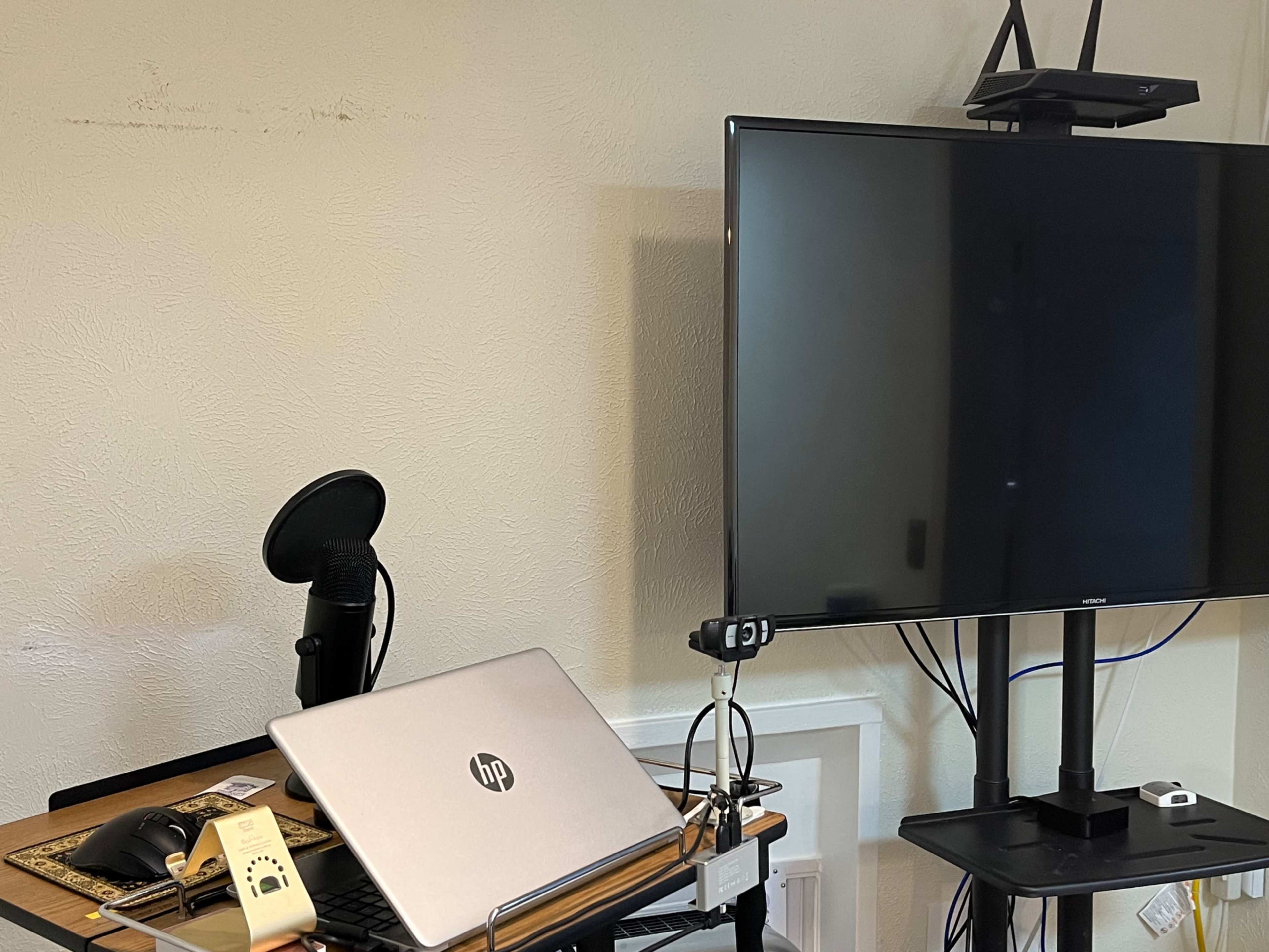 A silver HP laptop sits on a desk beside a microphone and a large wall-mounted television.