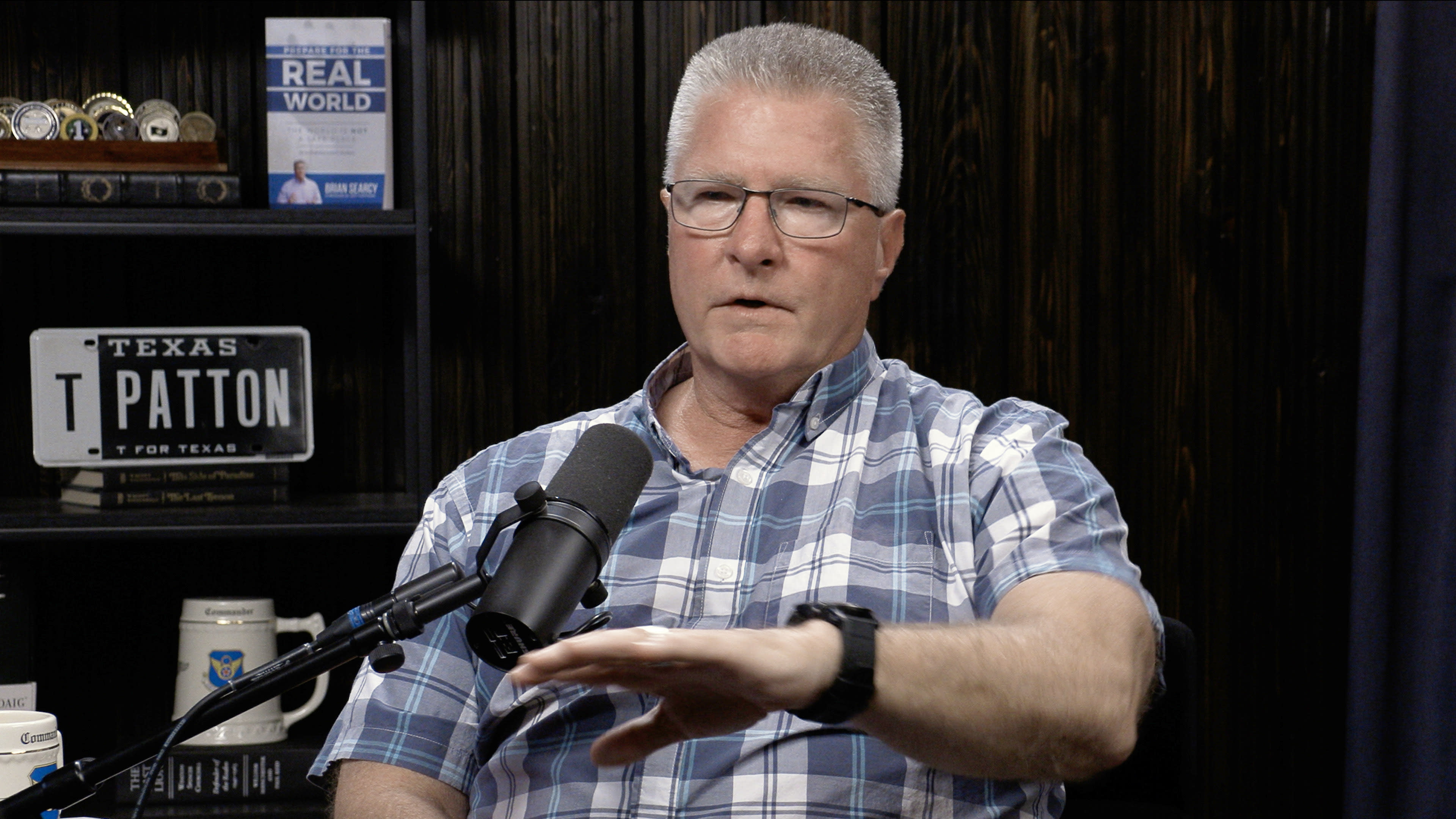 A man in a plaid shirt is speaking into a microphone while sitting in front of a bookshelf with various items, including a "Texas T for Texas" sign.