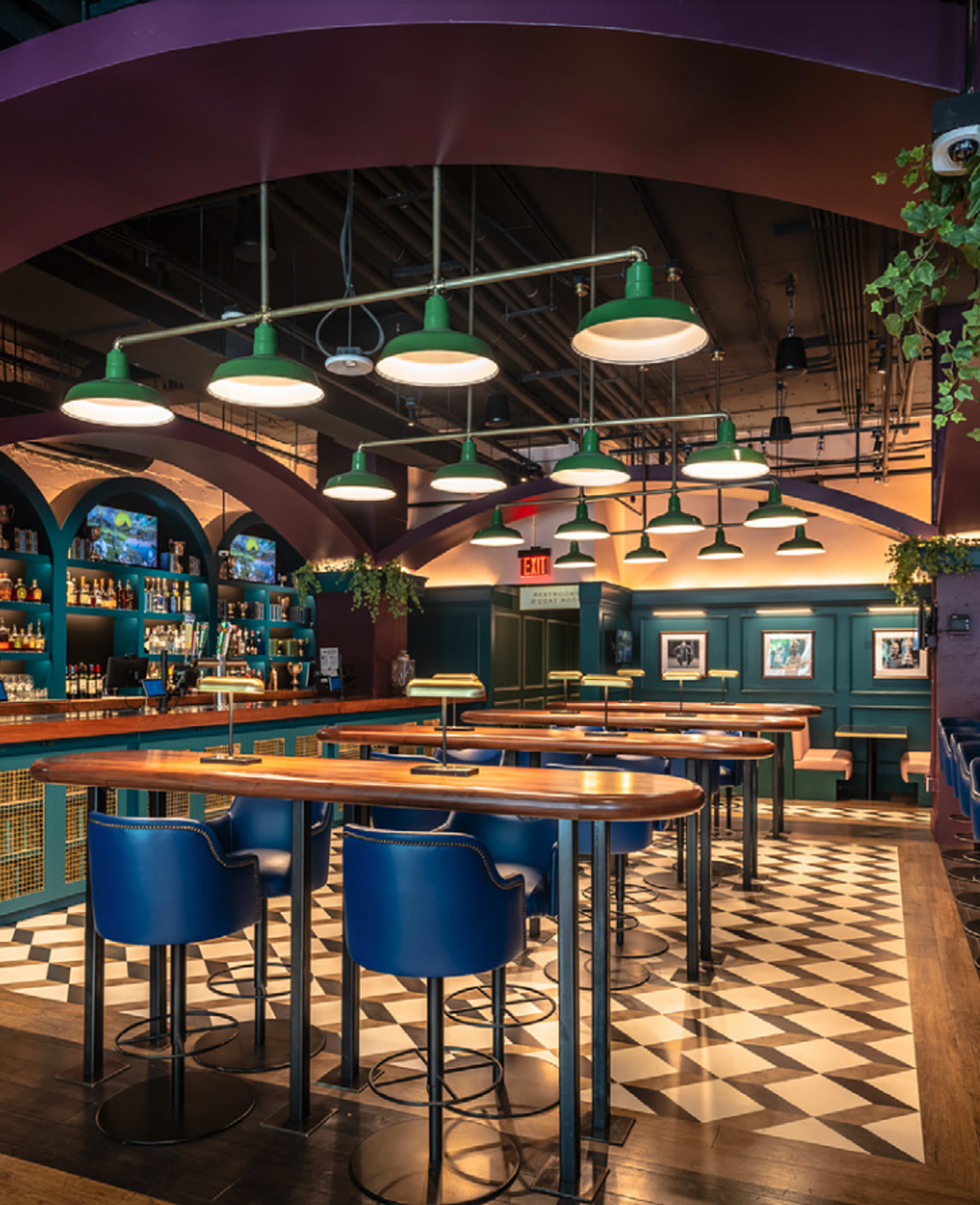 The image shows an upscale bar with a long wooden counter, blue bar stools, and bright green pendant lights, set against a backdrop of intricate tile flooring and dark blue walls lined with shelves of bottles.