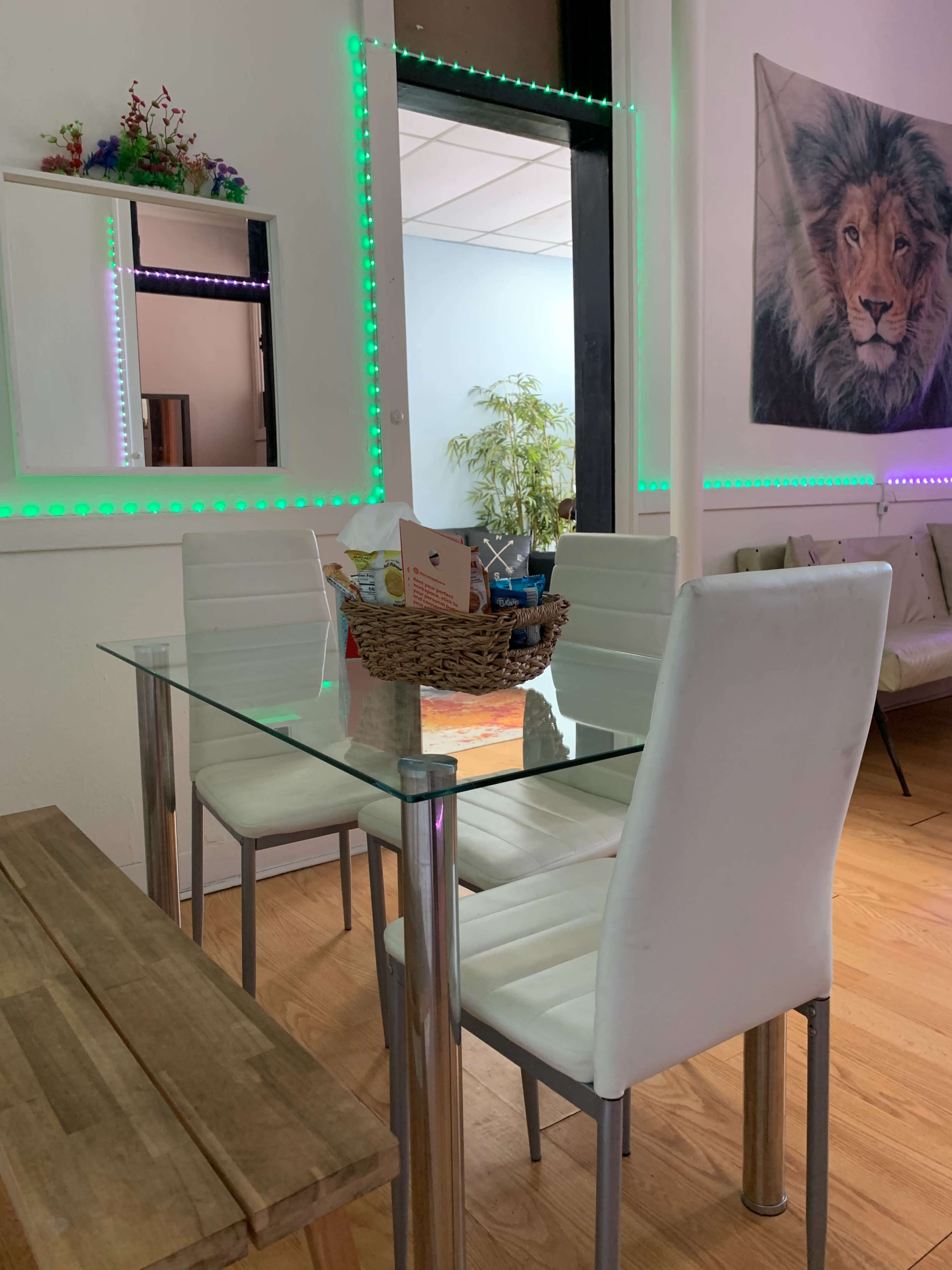 A glass dining table with white chairs is set in a room featuring a mirror and a lion-themed wall decoration, accompanied by LED lights around the mirror.
