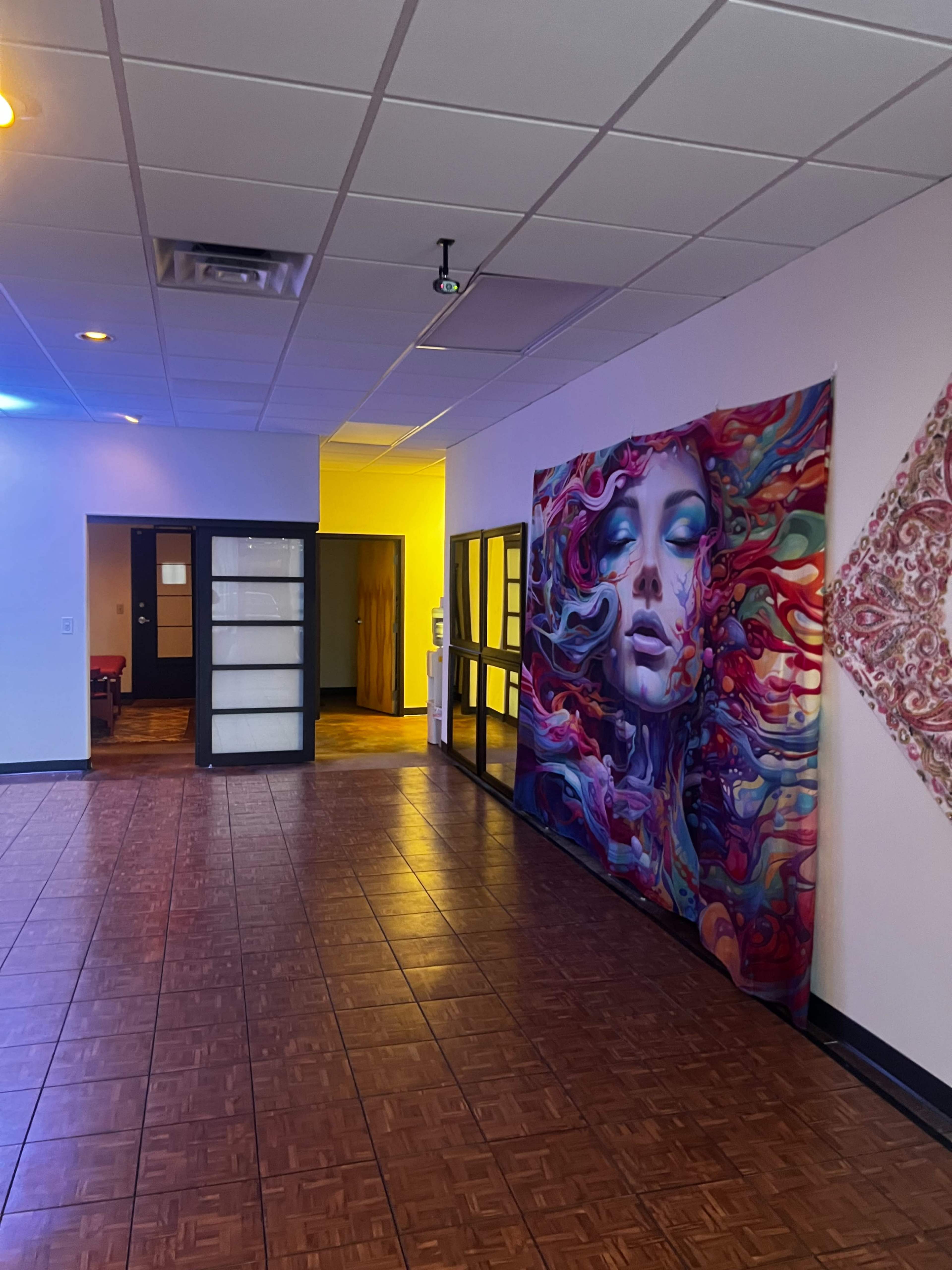The hallway features a large, colorful mural of a woman's face on one wall, with doors and a yellow-lit room in the background.