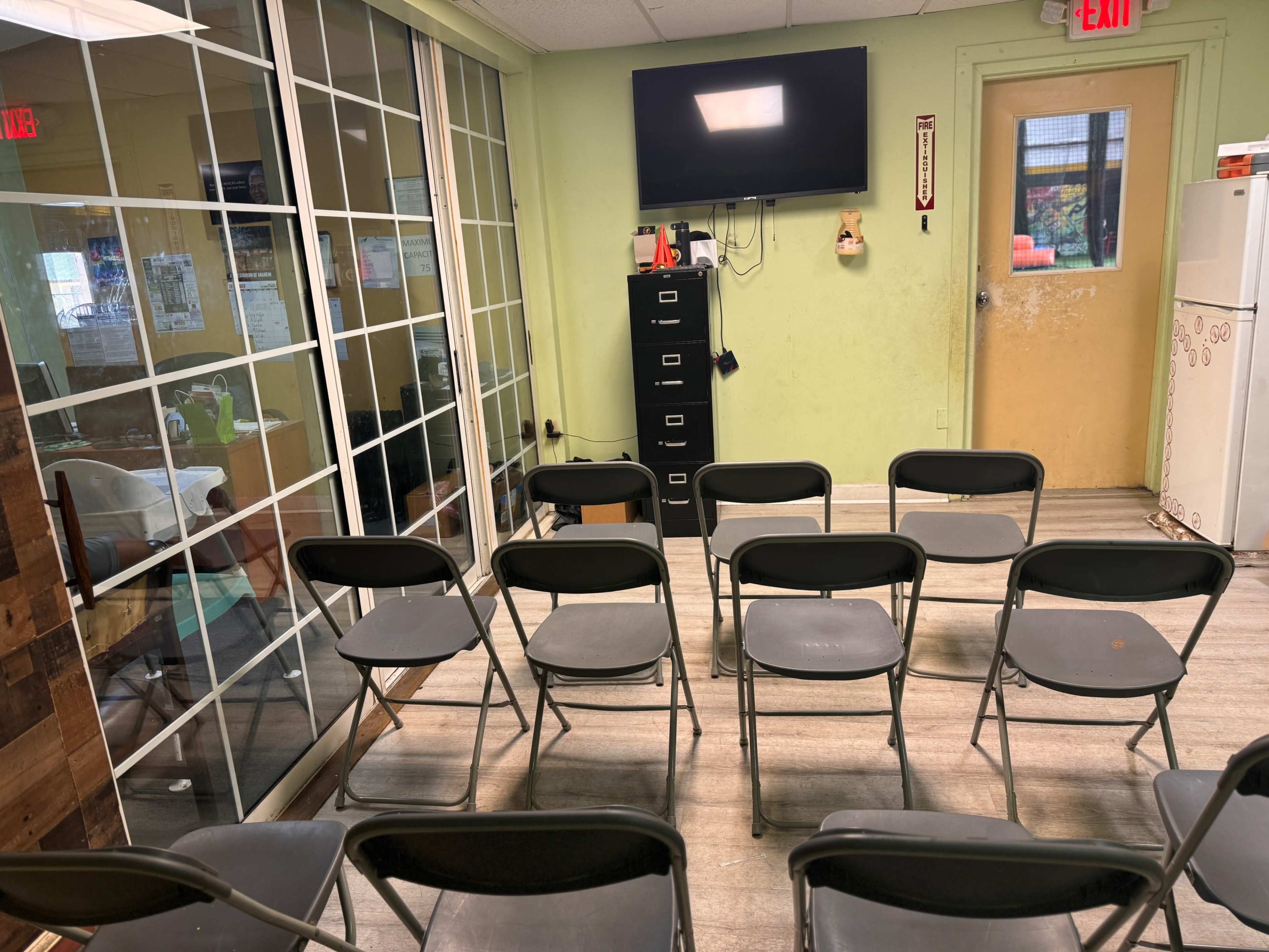 A room with several black folding chairs arranged in front of a wall-mounted television and a filing cabinet, with a door leading to another area visible in the background.