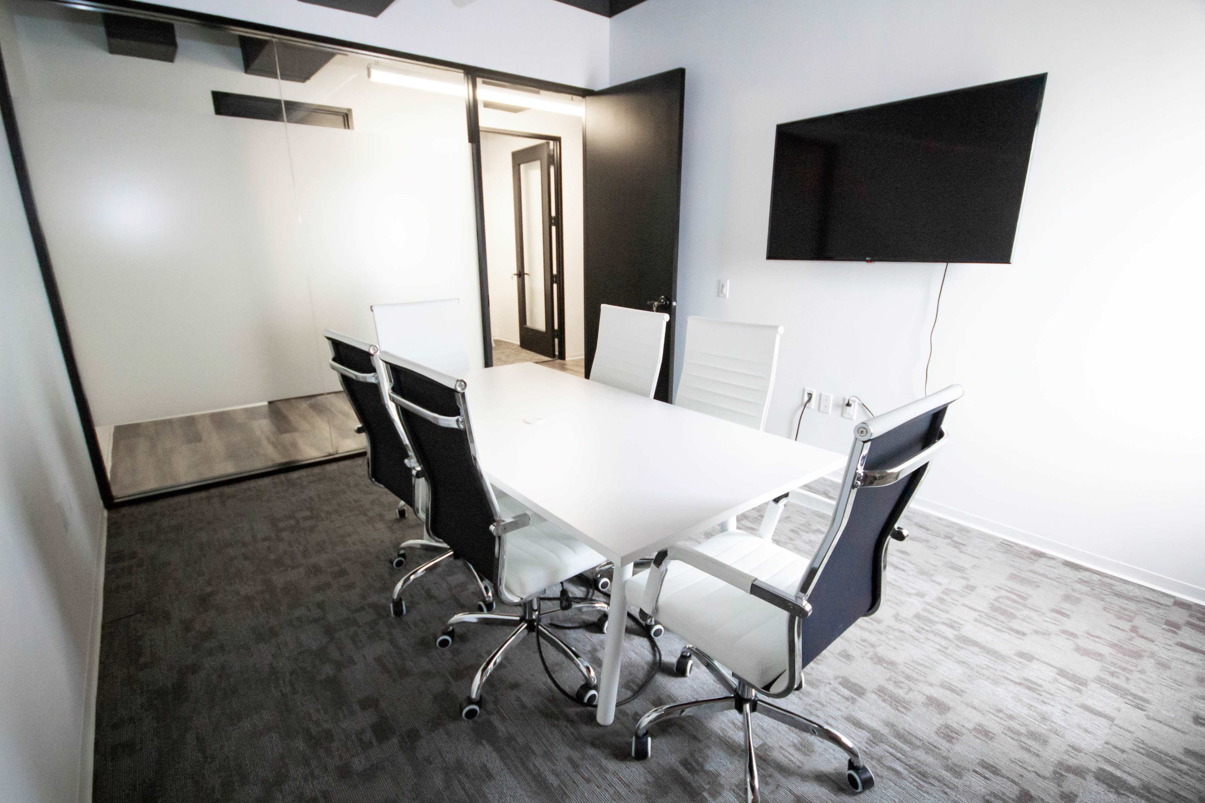 A modern conference room with a white table and six chairs, a large screen mounted on the wall, and a glass wall separating it from an adjacent space.