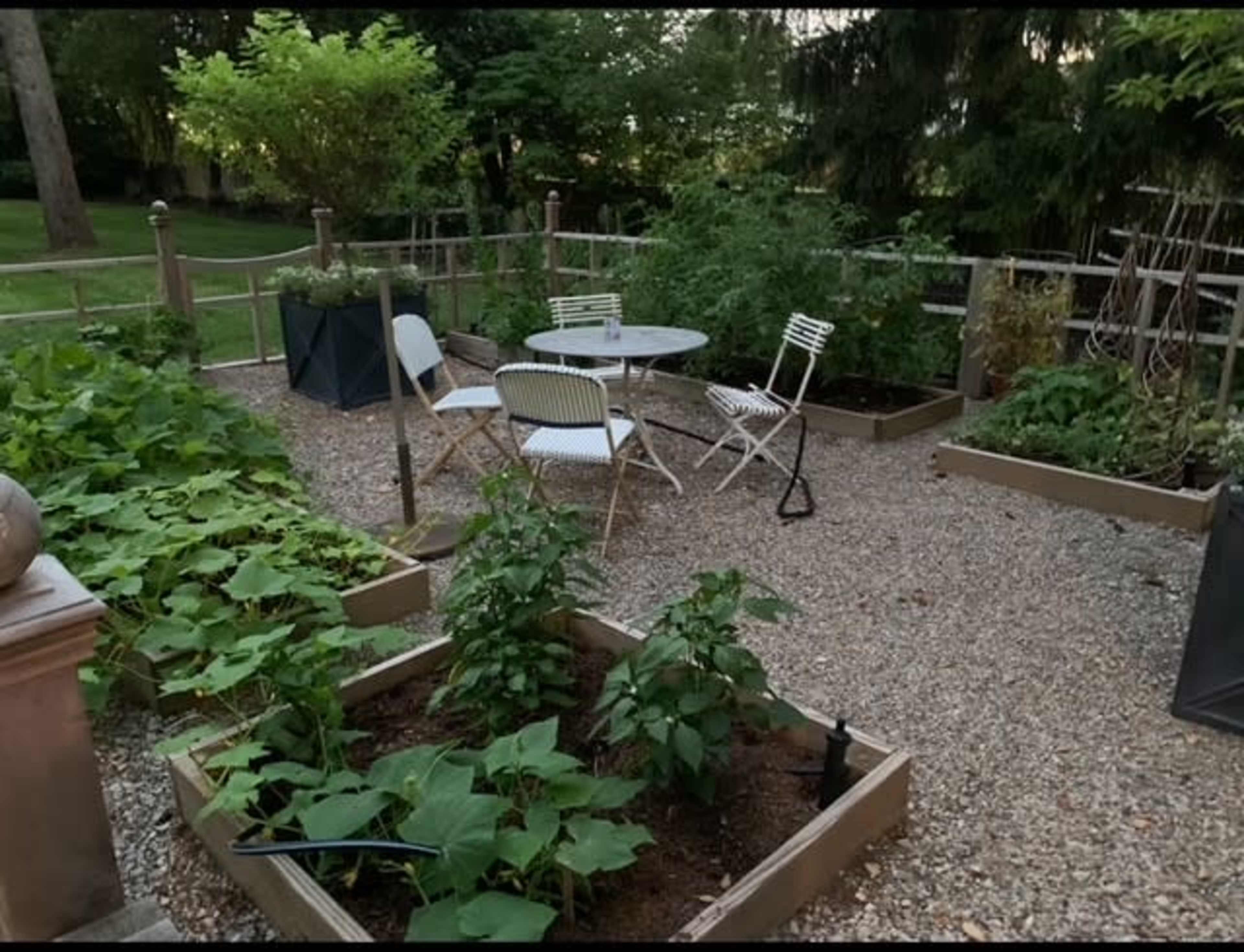 The image shows a garden patio area with raised planting beds containing vegetables, a small table and chairs, and greenery in the background.
