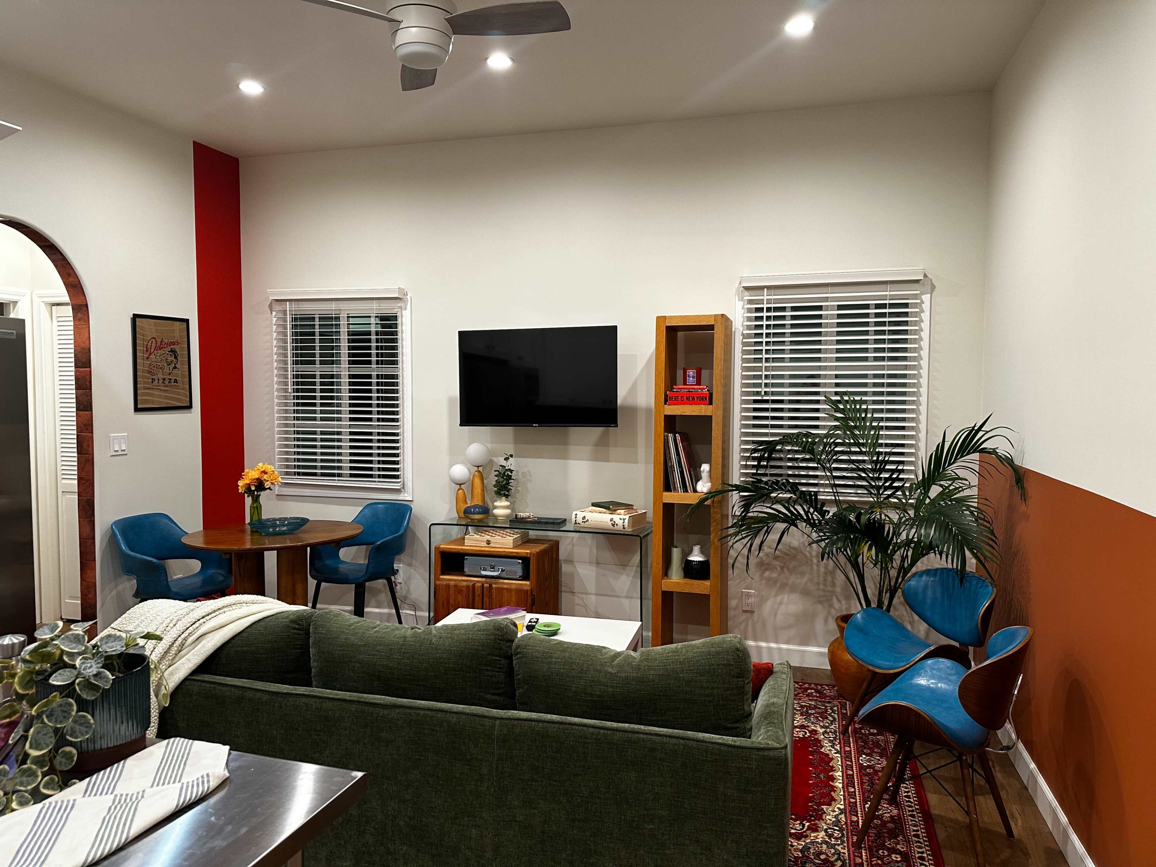 The image shows a cozy living room with a green sofa, a wooden coffee table, two blue chairs, a plant, and a wall-mounted TV above a small wooden shelf.