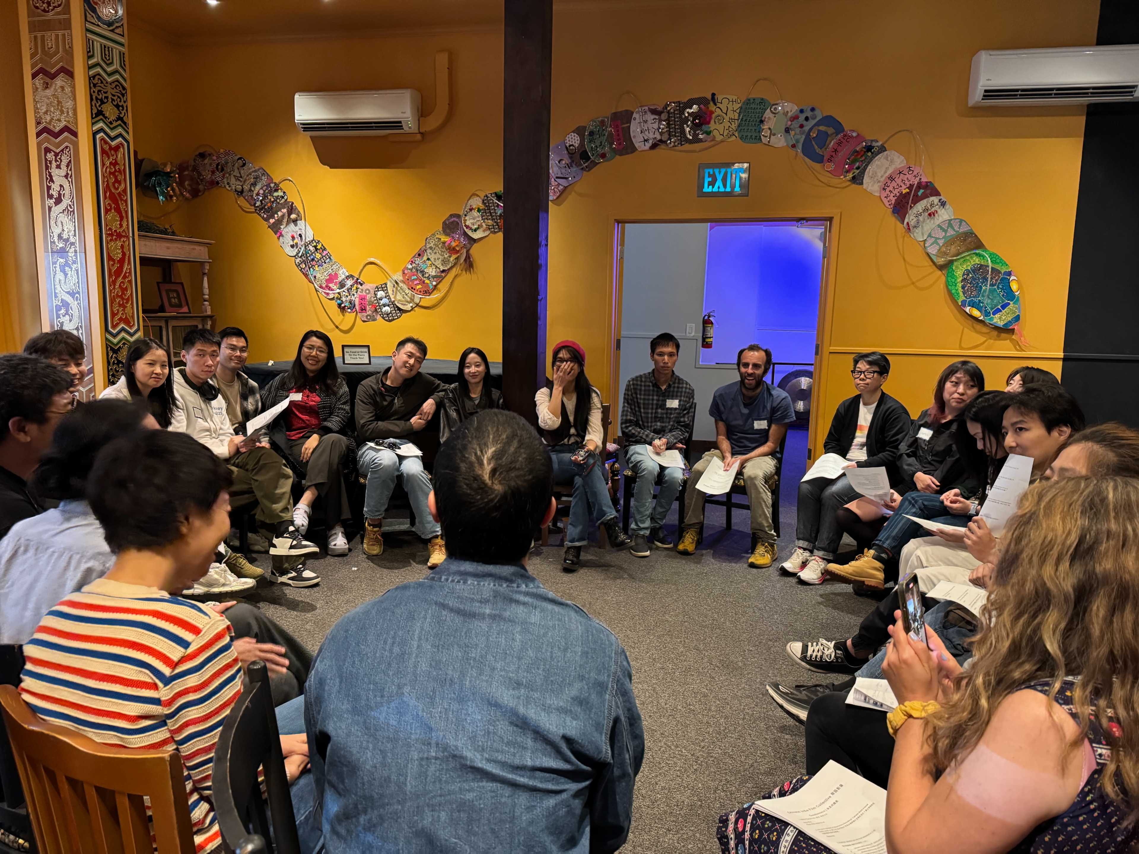 A diverse group of people sits in a circular arrangement in a brightly colored room, engaging in discussion or a meeting.