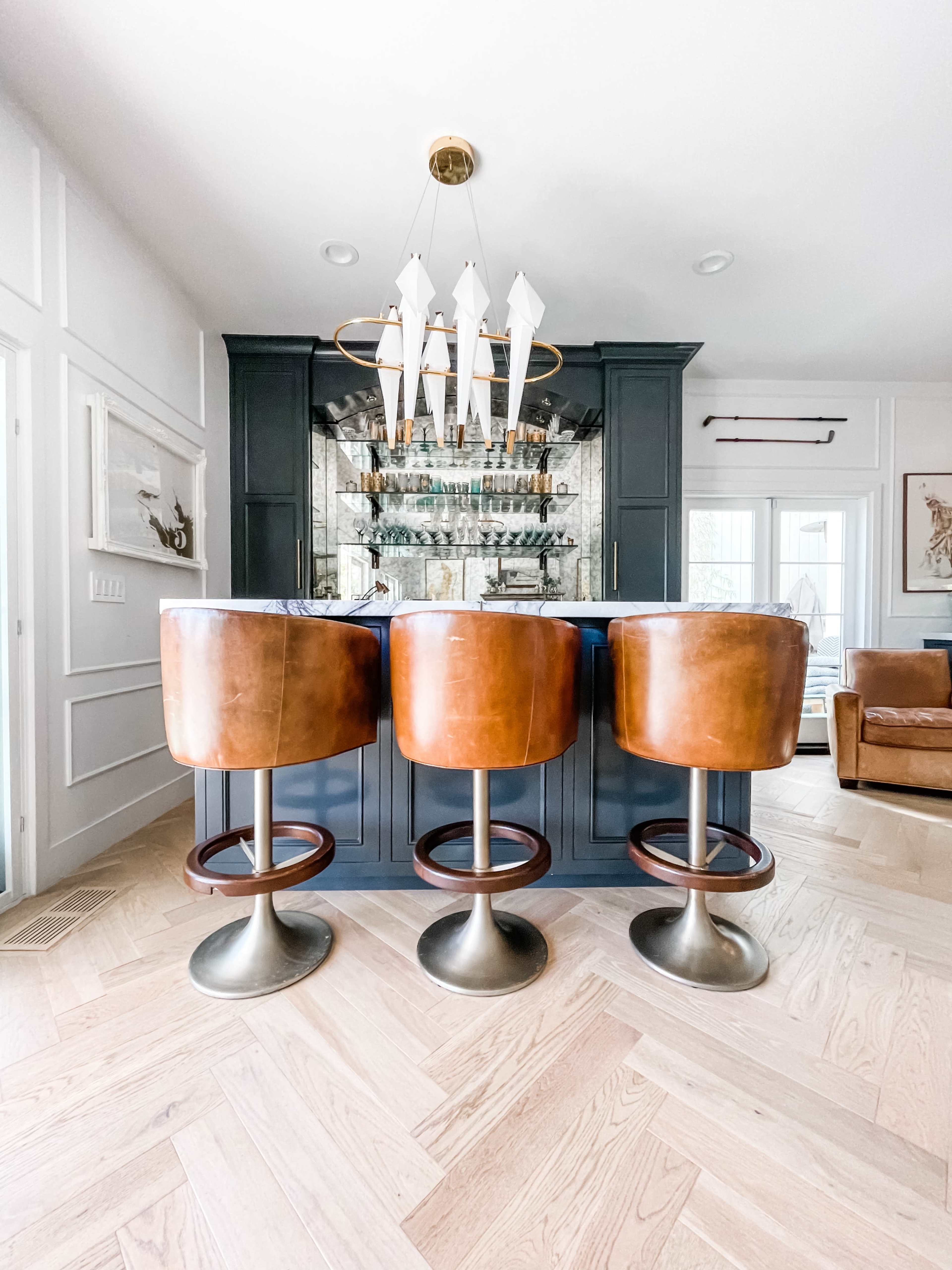 A modern bar area features three brown leather swivel stools in front of a stylish, dark wood bar with a glass display of bottles and a contemporary light fixture overhead.