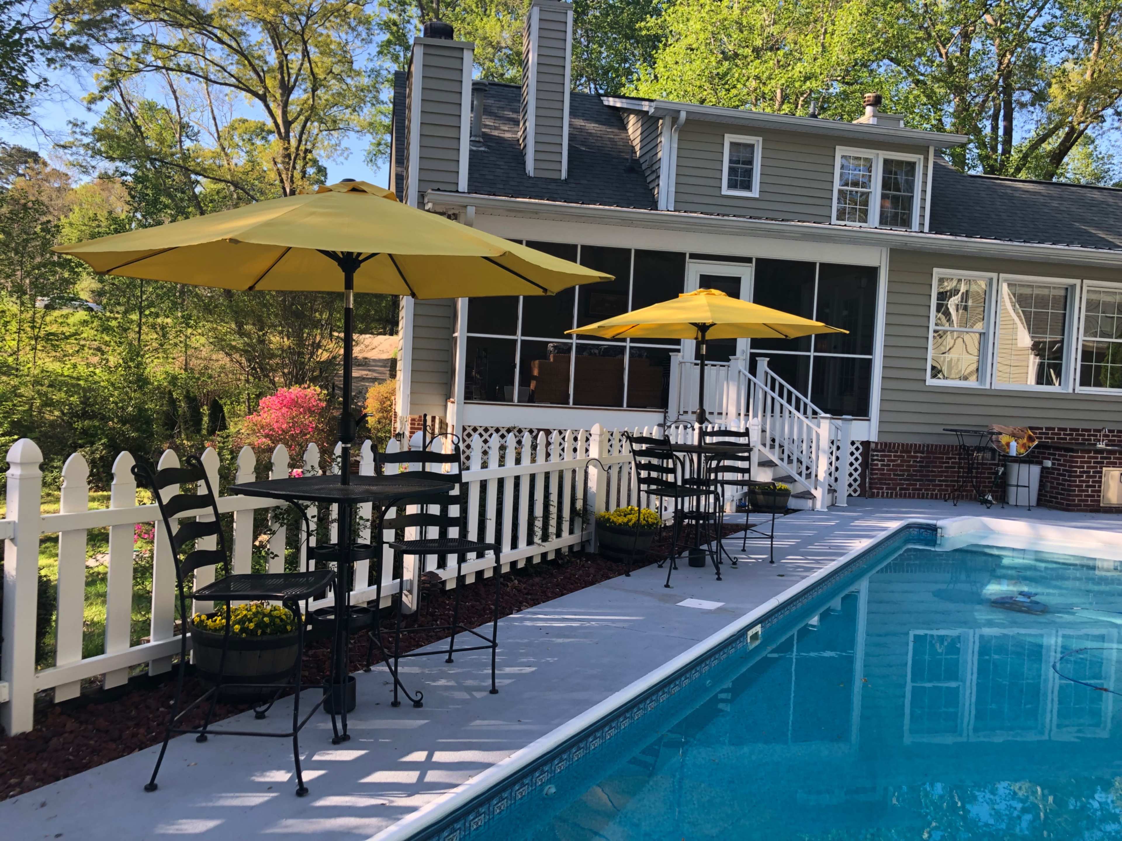A backyard pool area features two yellow umbrellas over black metal tables beside the water, with a fenced garden in the background.