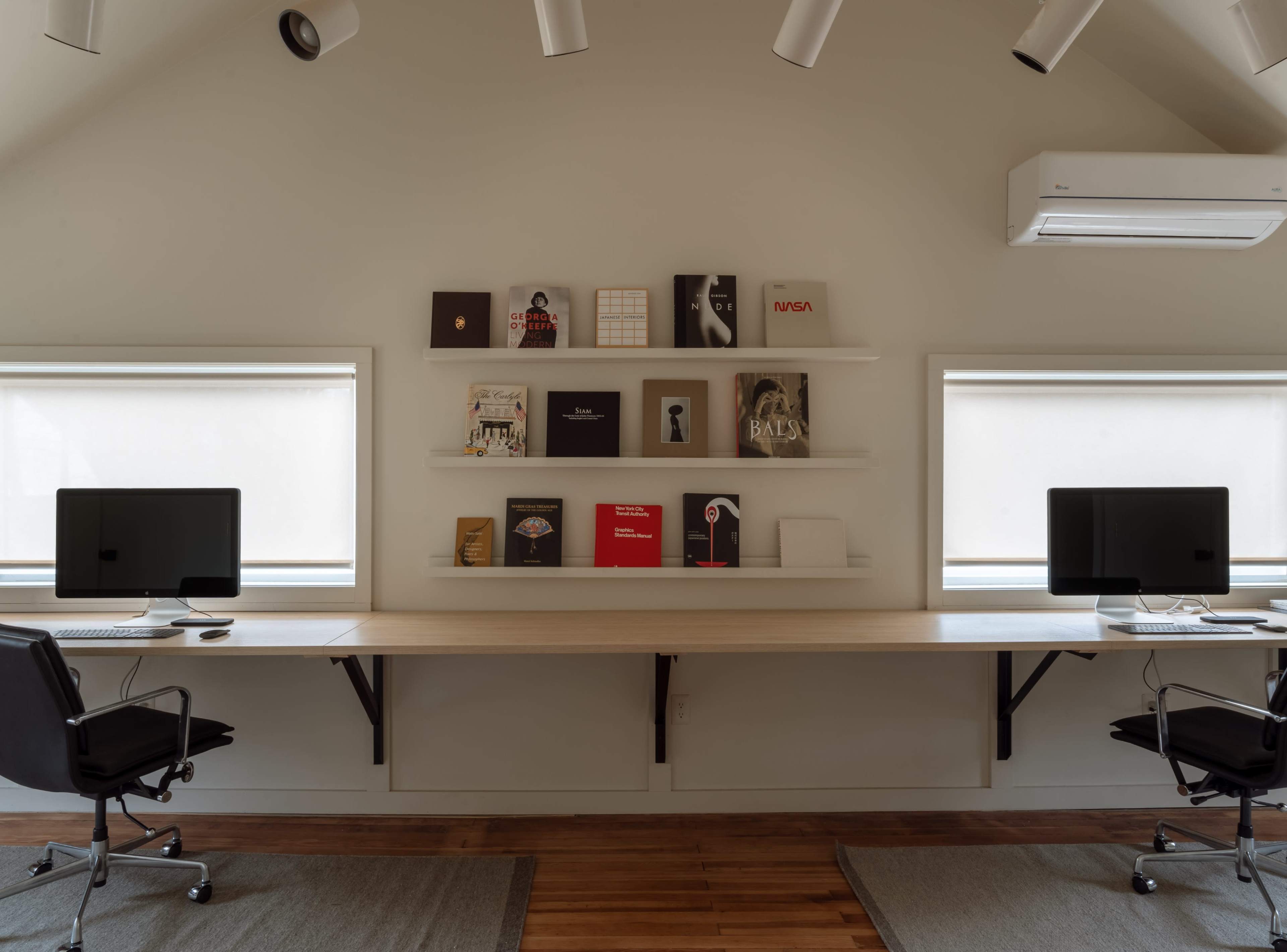 A modern workspace with two computers on a long wooden desk, flanked by shelves displaying various books and magazines above, along with two large windows.