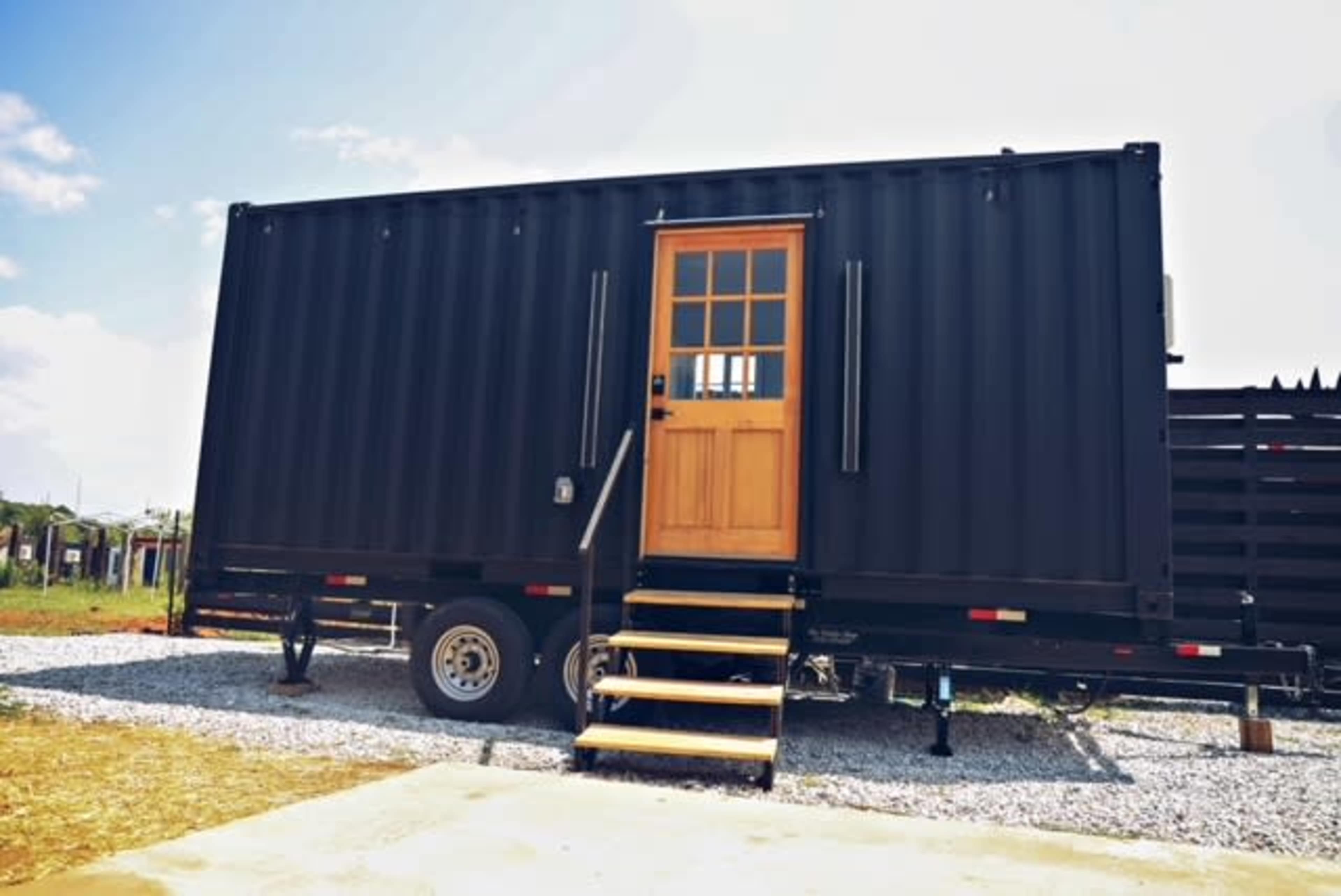 A black shipping container with a wooden door and steps sits on a gravel surface, surrounded by an open landscape.
