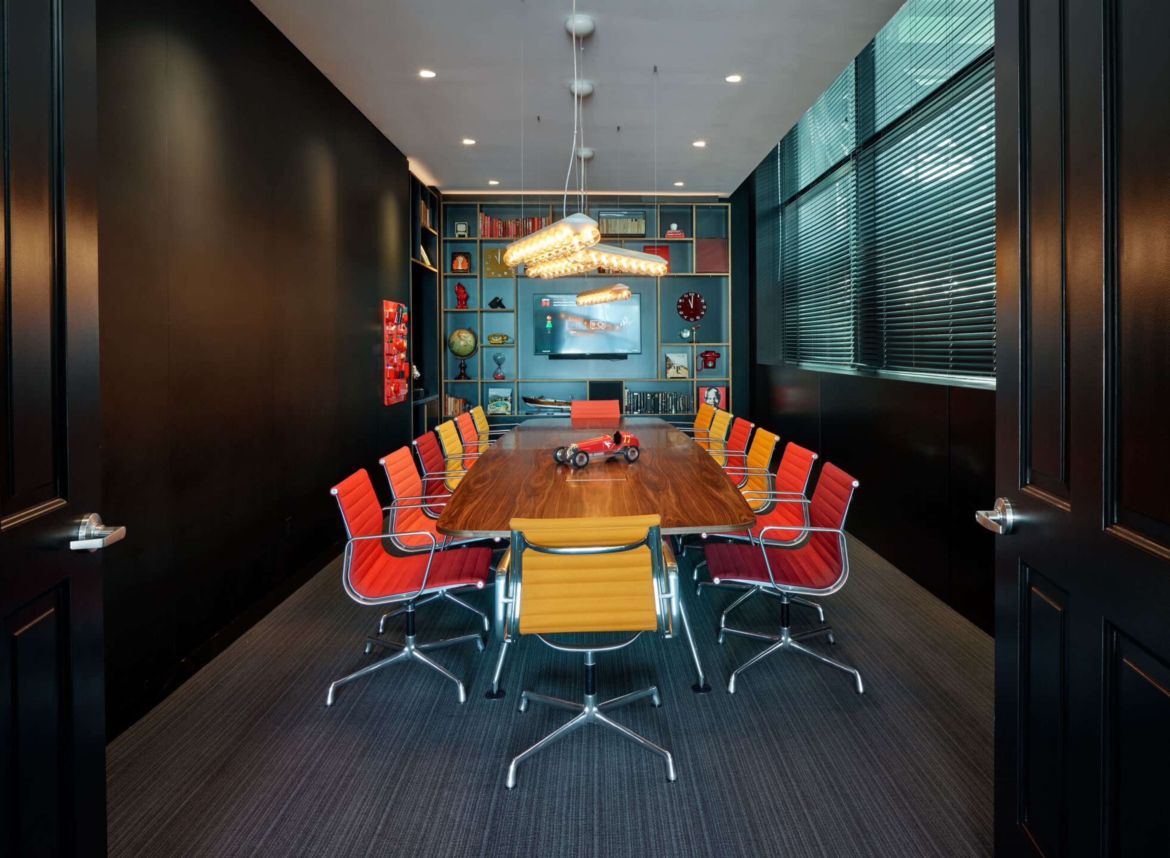 A modern conference room features a long wooden table surrounded by colorful chairs, with shelves displaying decorative items in the background.