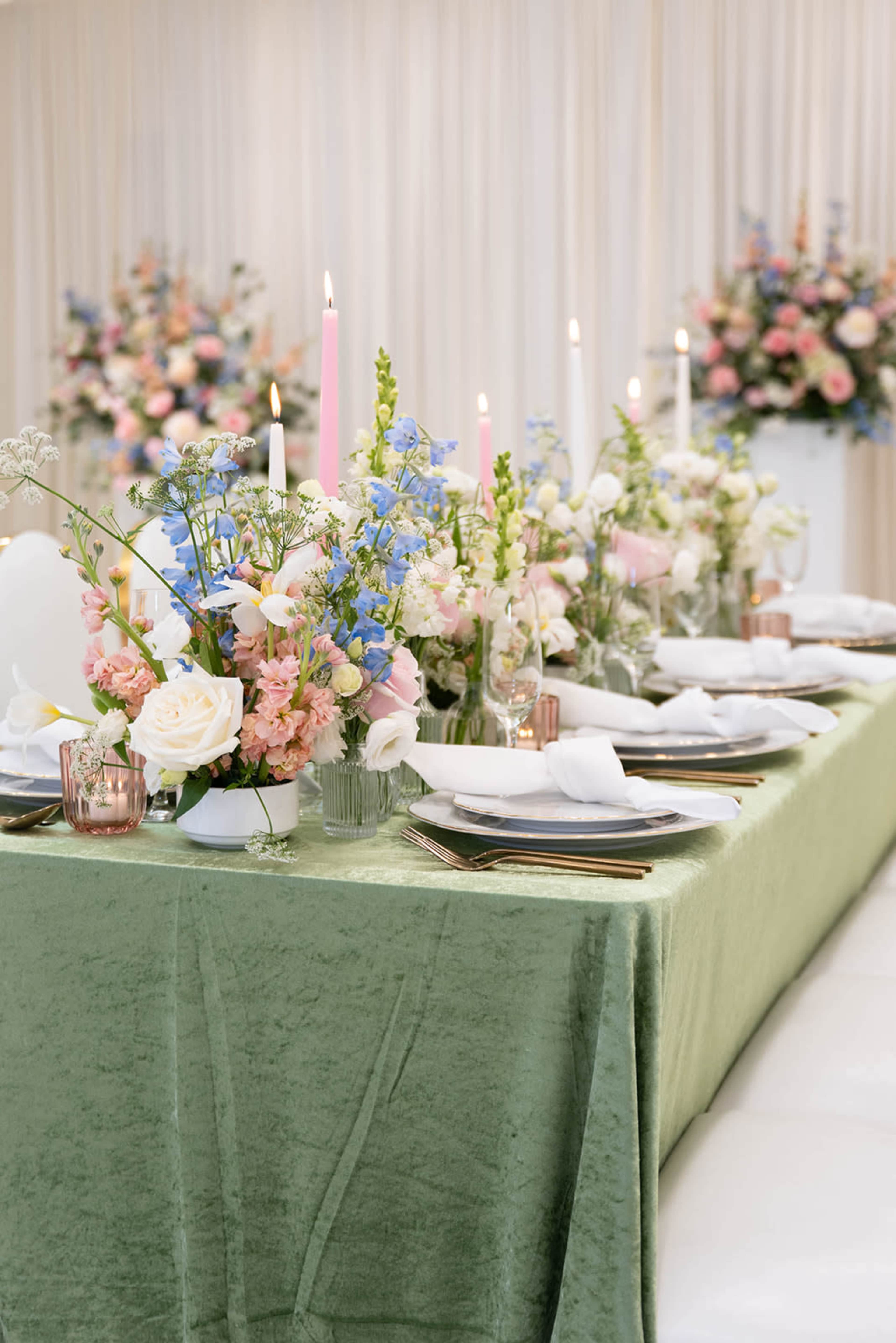 The image shows a beautifully set dining table with a green tablecloth, adorned with floral centerpieces and pink candles, surrounded by white seating.