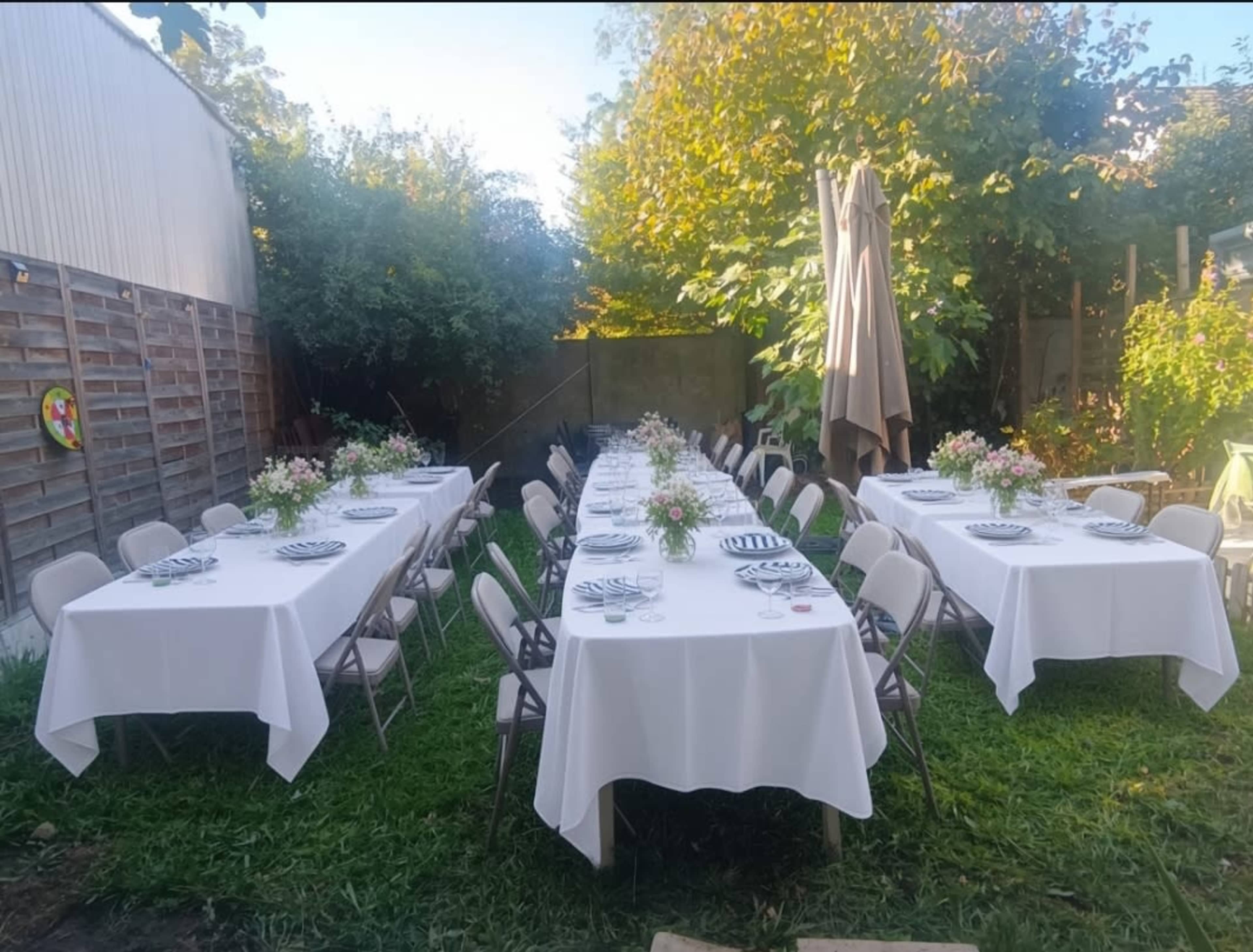 A long table is set with white tablecloths and decorative flower arrangements in a garden area, surrounded by greenery.