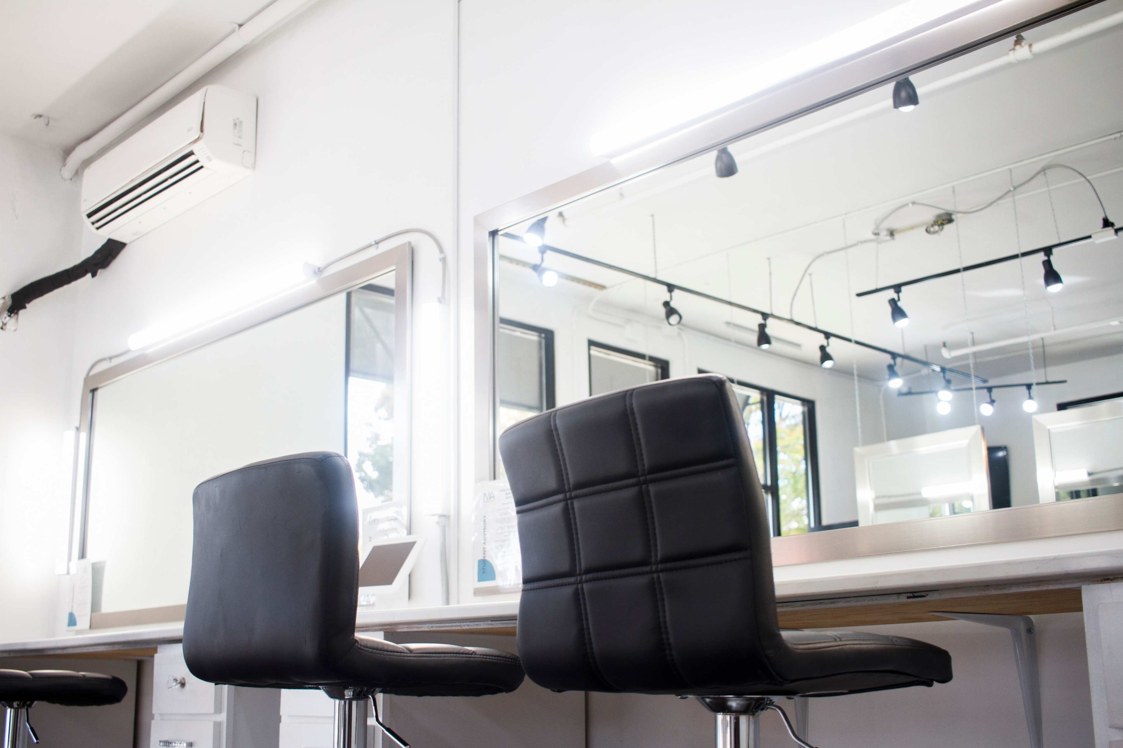 Two black salon chairs facing a large mirror with bright overhead lighting in a minimalist salon interior.